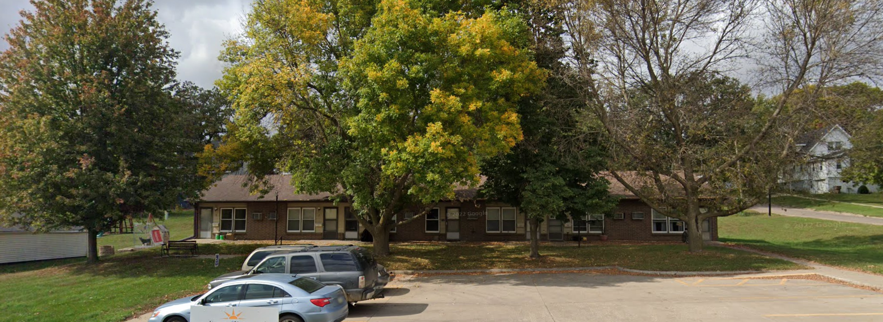 Parking lot with three parked cars and trees with green and yellow leaves in front of a brown brick building and a white house in the background.