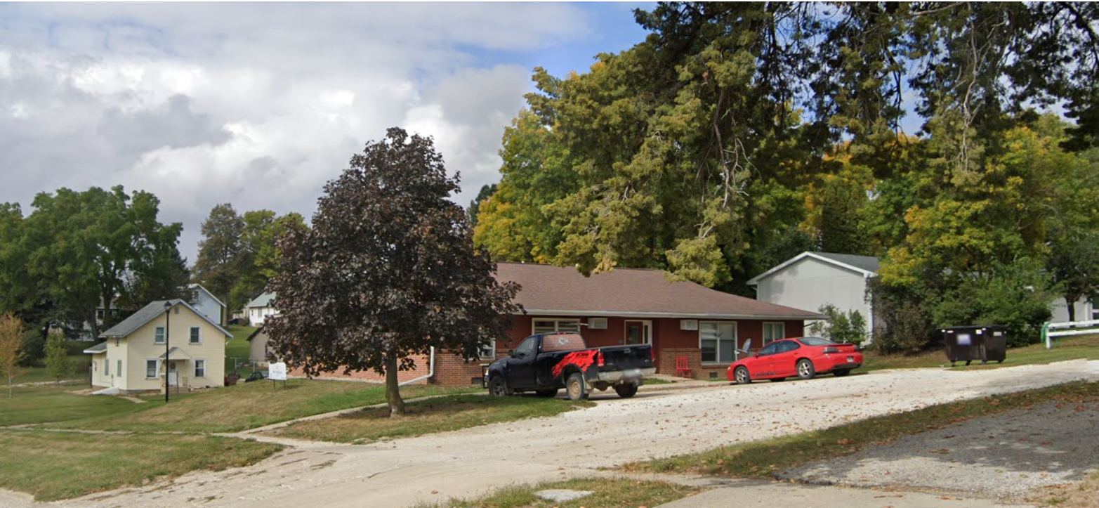 A residential neighborhood with a brick house, parked vehicles including a black pickup truck and a red car, a large tree in front, and other houses in the background under partly cloudy skies.
