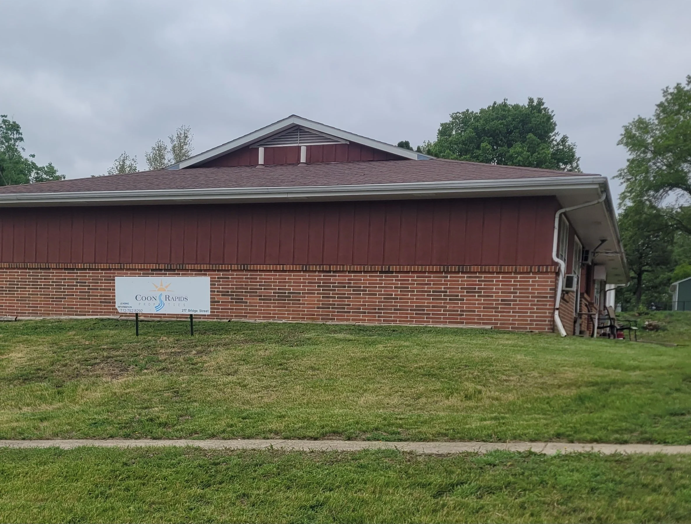 A two-story house with a brick lower section and a reddish-brown upper section, topped with a gabled roof. There is a sign in front of the house, and it is on a grassy lawn with some trees in the background and an overcast sky.