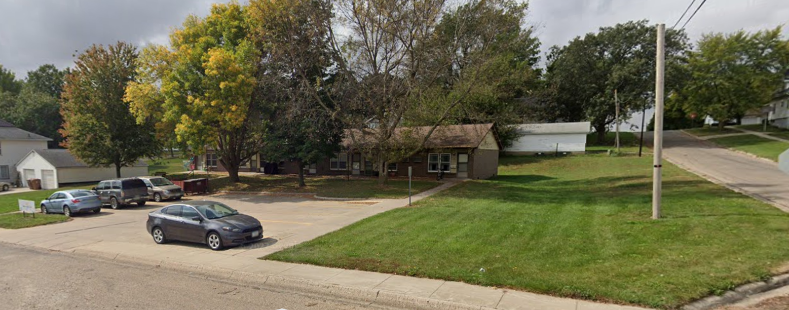 A grassy area with trees and houses in the background, a parking lot with several cars, and a utility pole with wires.
