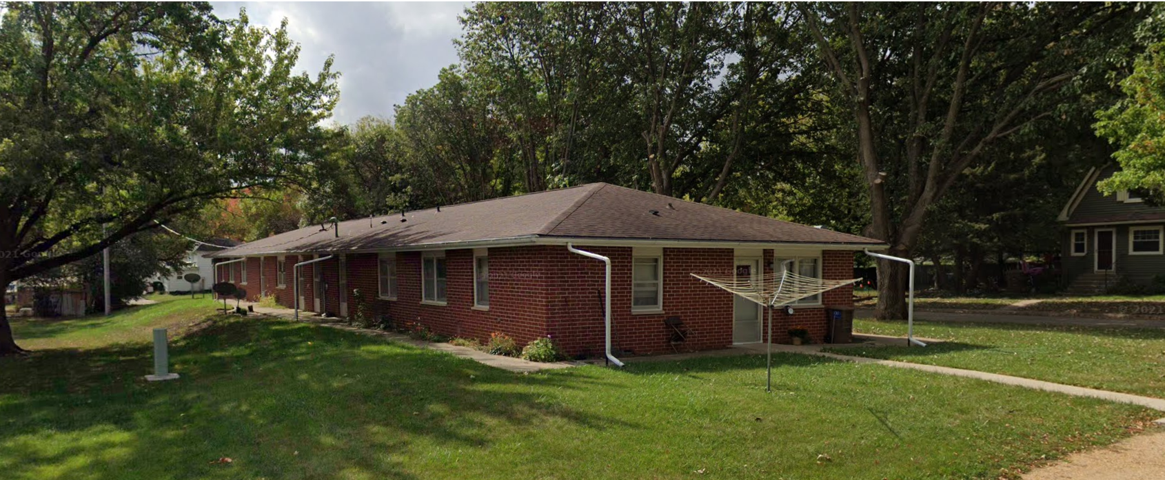A single-story brick house with a brown shingle roof, surrounded by green trees and lawn, with a clothesline in the yard.