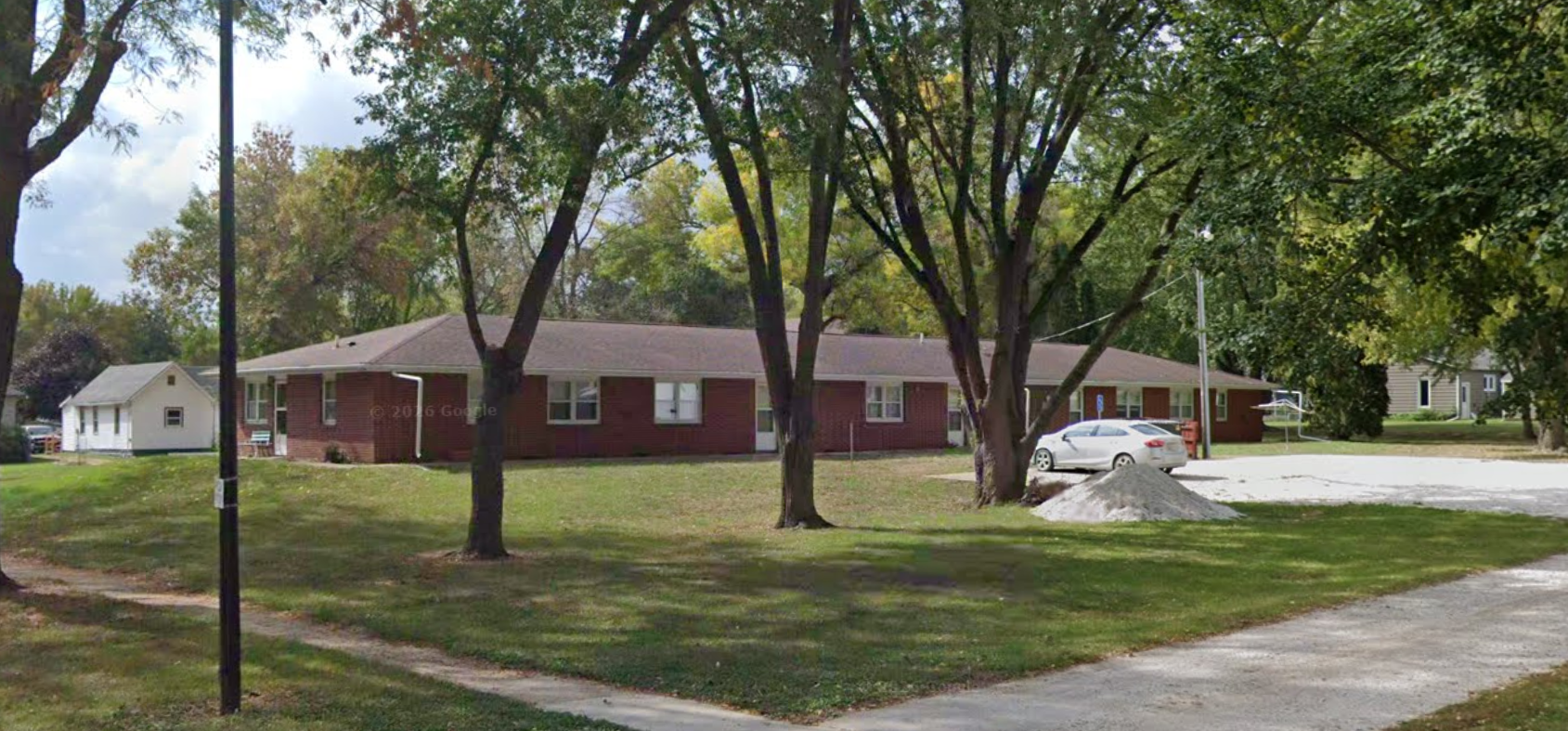 A residential scene with a single-story brick house, several trees, a parked white car, and a gravel driveway on a cloudy day.