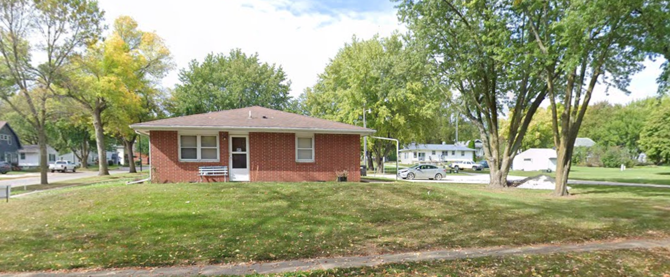 A small red brick building with a white door and two windows, surrounded by green grass and tall trees in a neighborhood.