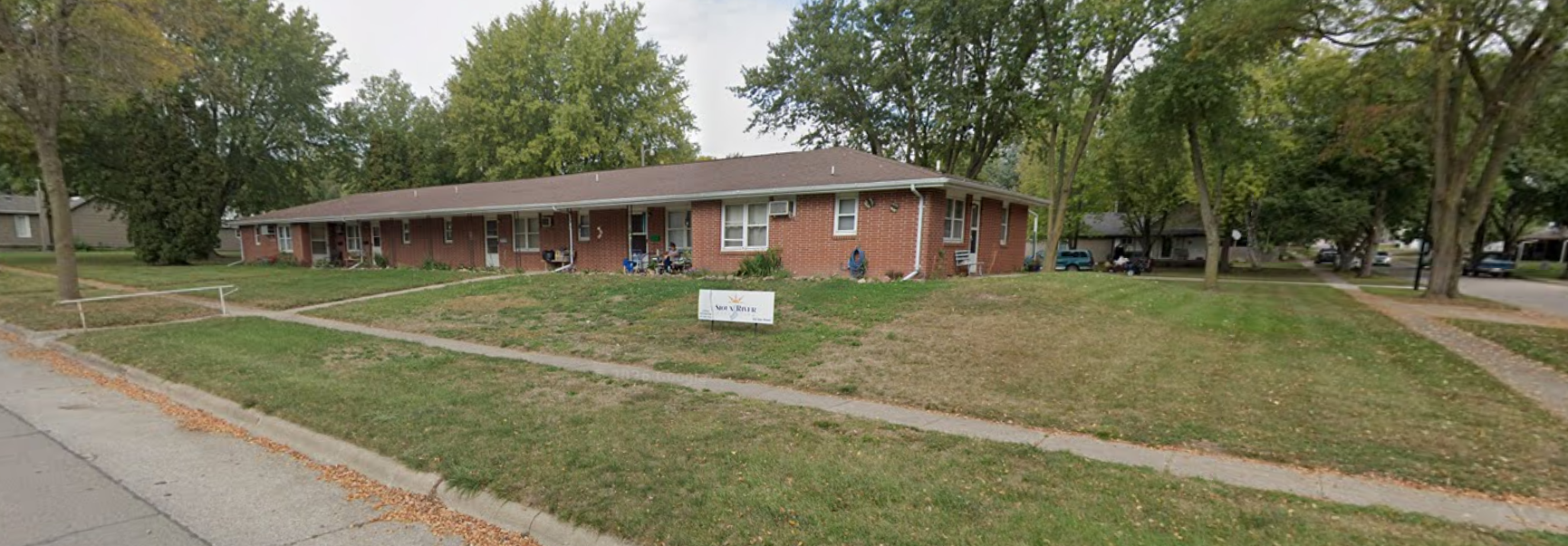 A single-story brick building with multiple apartment entrances, surrounded by trees and a grassy yard with walkways.
