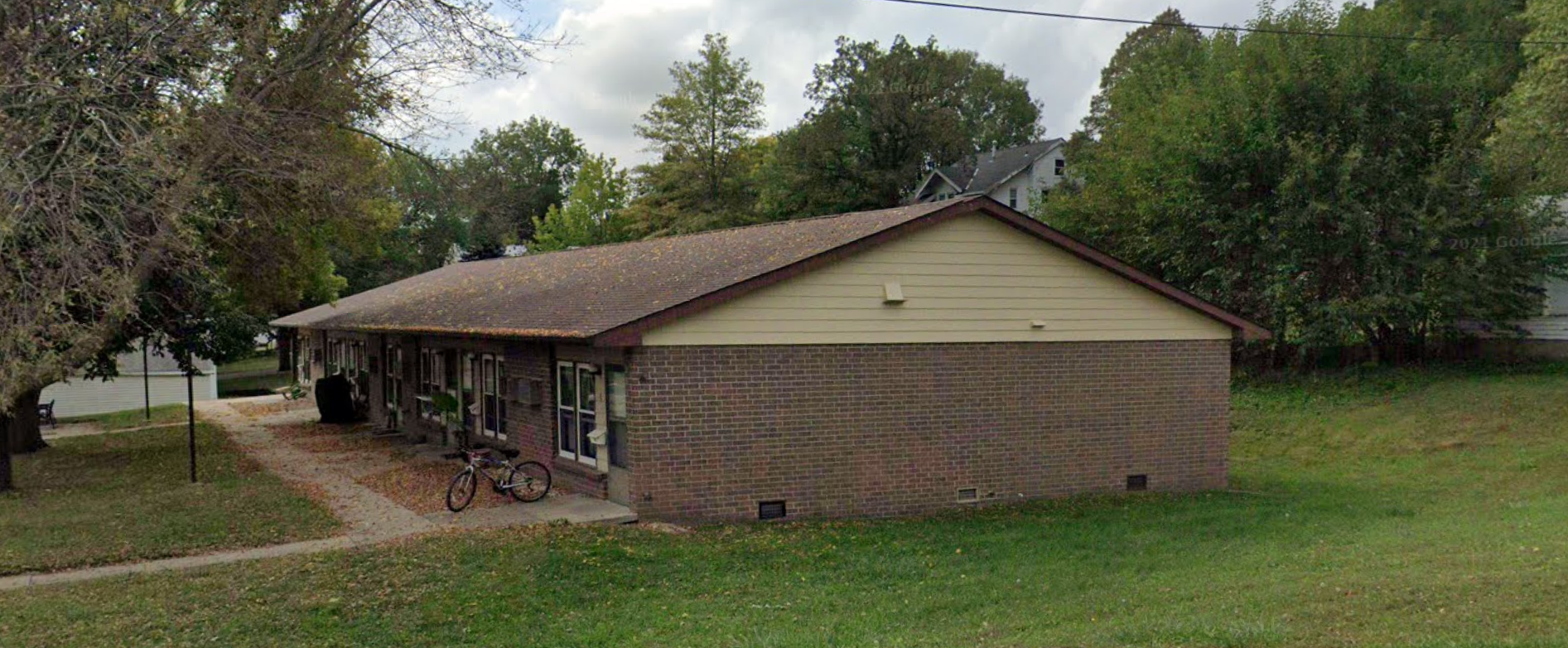 A single-story brick house with a sloped shingle roof, surrounded by trees and green grass, with a bicycle parked outside near the front entrance.