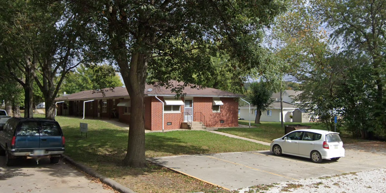 A small brick apartment complex with a front lawn, surrounded by trees, with a parking lot containing a few cars.