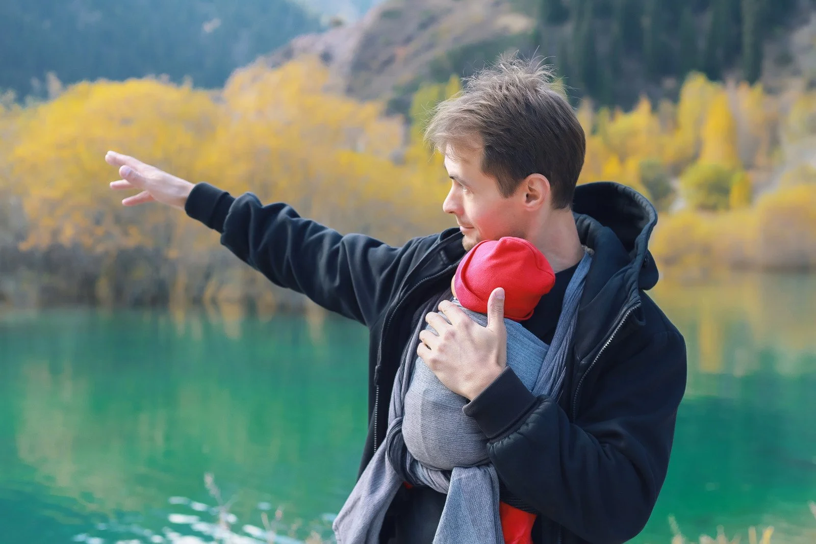 A young man is holding a baby in a baby carrier outdoors near a body of water with a forested area with yellow autumn foliage in the background. The young man is pointing with his right arm, and the baby's face is not visible as it is turned away.