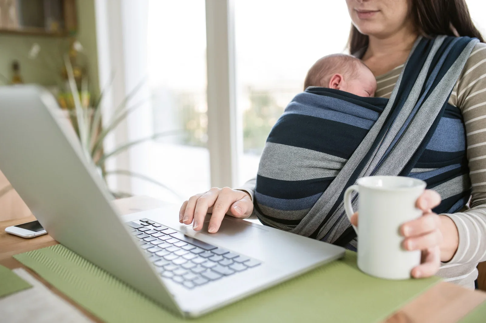 Woman working at a laptop with a baby in a front carrier, holding a mug.