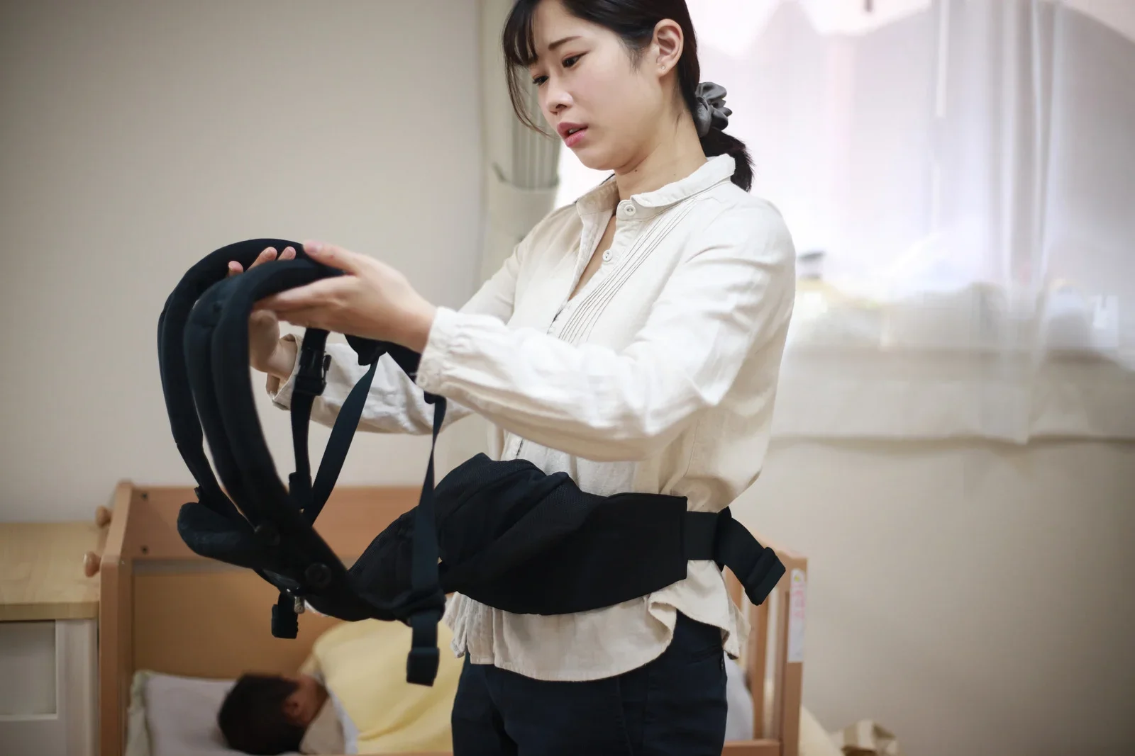 A woman with dark hair tied back and wearing a cream-colored blouse is standing in a bedroom, looking at and adjusting a black harness or brace, with a person lying on a bed in the background.