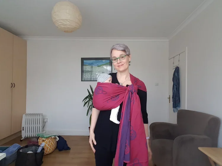 A woman with short gray hair, glasses, and a black outfit carrying a baby wrapped in a maroon sling, standing in a living room with a beige couch, a wardrobe, and a painting on the wall.