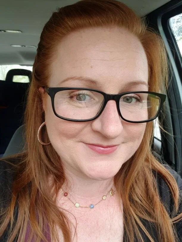 A woman with red hair and glasses smiling inside a vehicle.