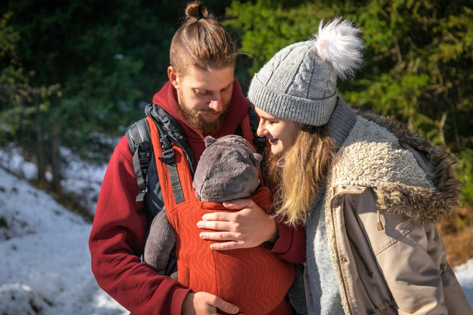 A family of three, a man, a woman, and a baby, outdoors in a snowy and wooded area. They are smiling and embracing each other, with the baby in the center.