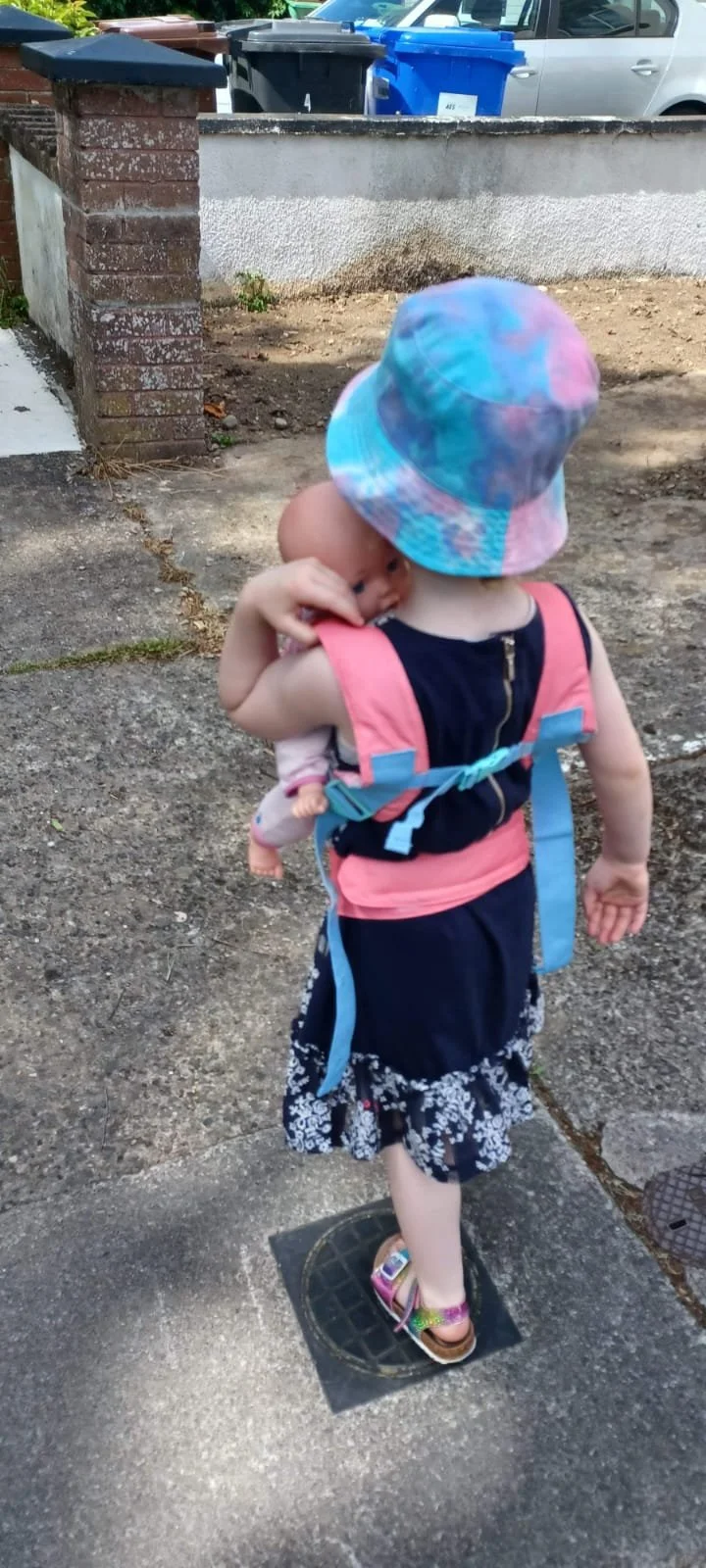 A young girl wearing a colorful bucket hat and a pink backpack holding a baby girl with a pink outfit and a pacifier, walking on a sidewalk with parking and trash bins in the background.