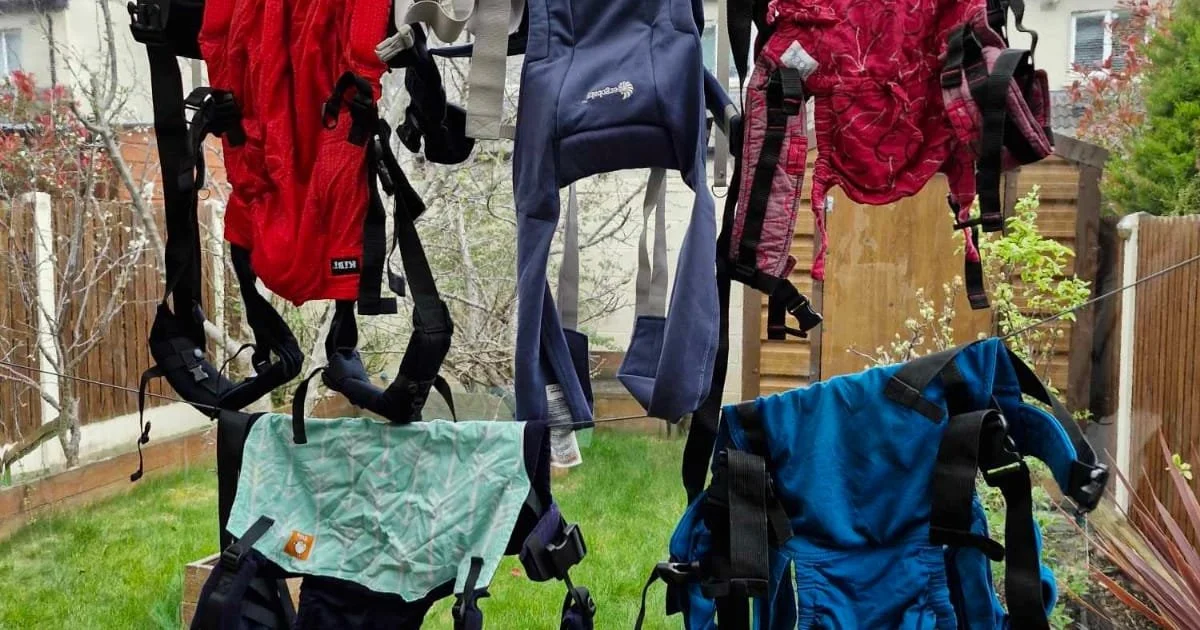 Clothes hanging on a makeshift outdoor clothesline, including backpacks, jackets, and pants, in a backyard with a wooden fence and trees.