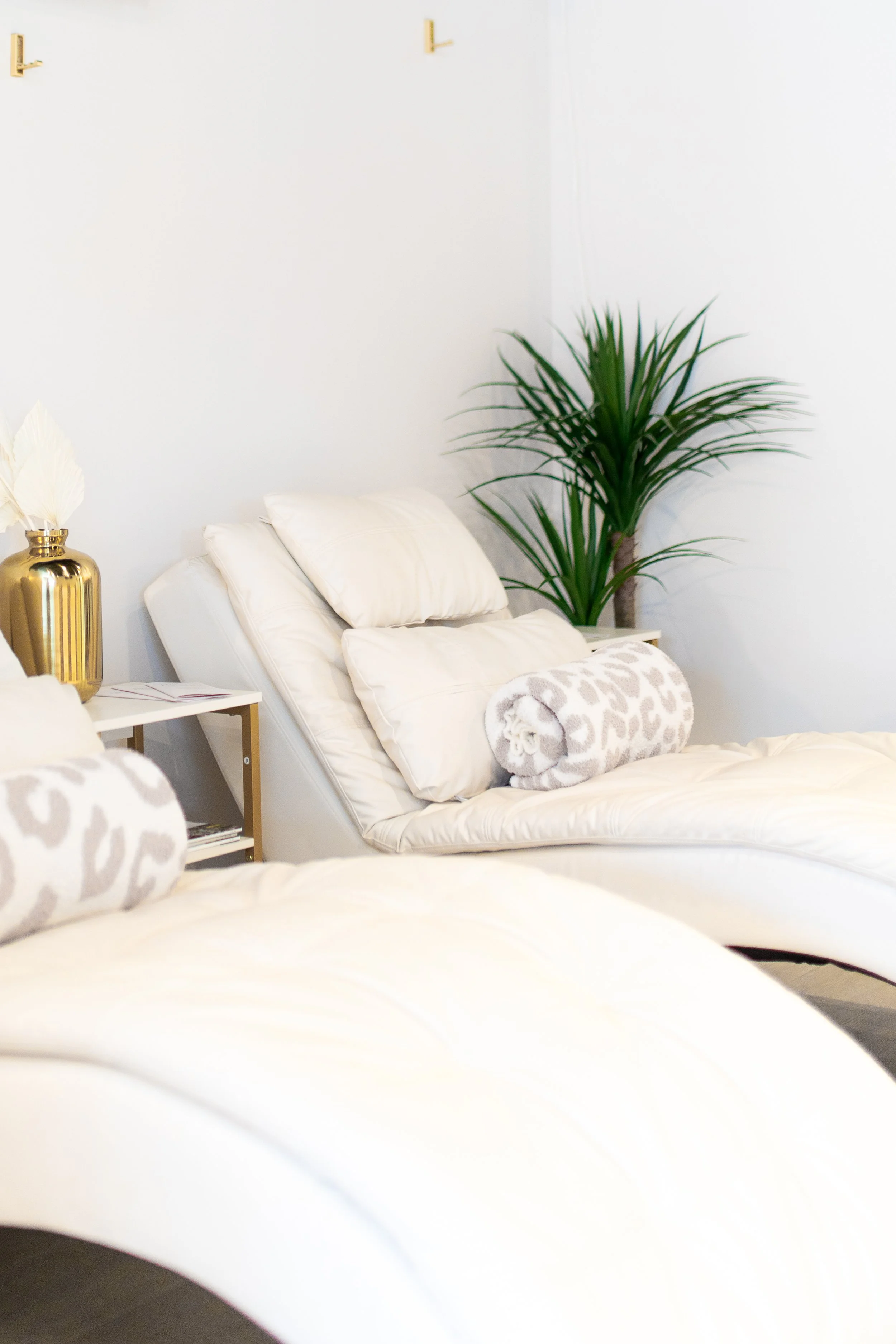 A bedroom with white bedding, pillows, and a rolled-up blanket. There is a gold side table with a decorative vase and plant on it, and a large green indoor plant next to the bed. The wall is white with minimalist gold hooks.