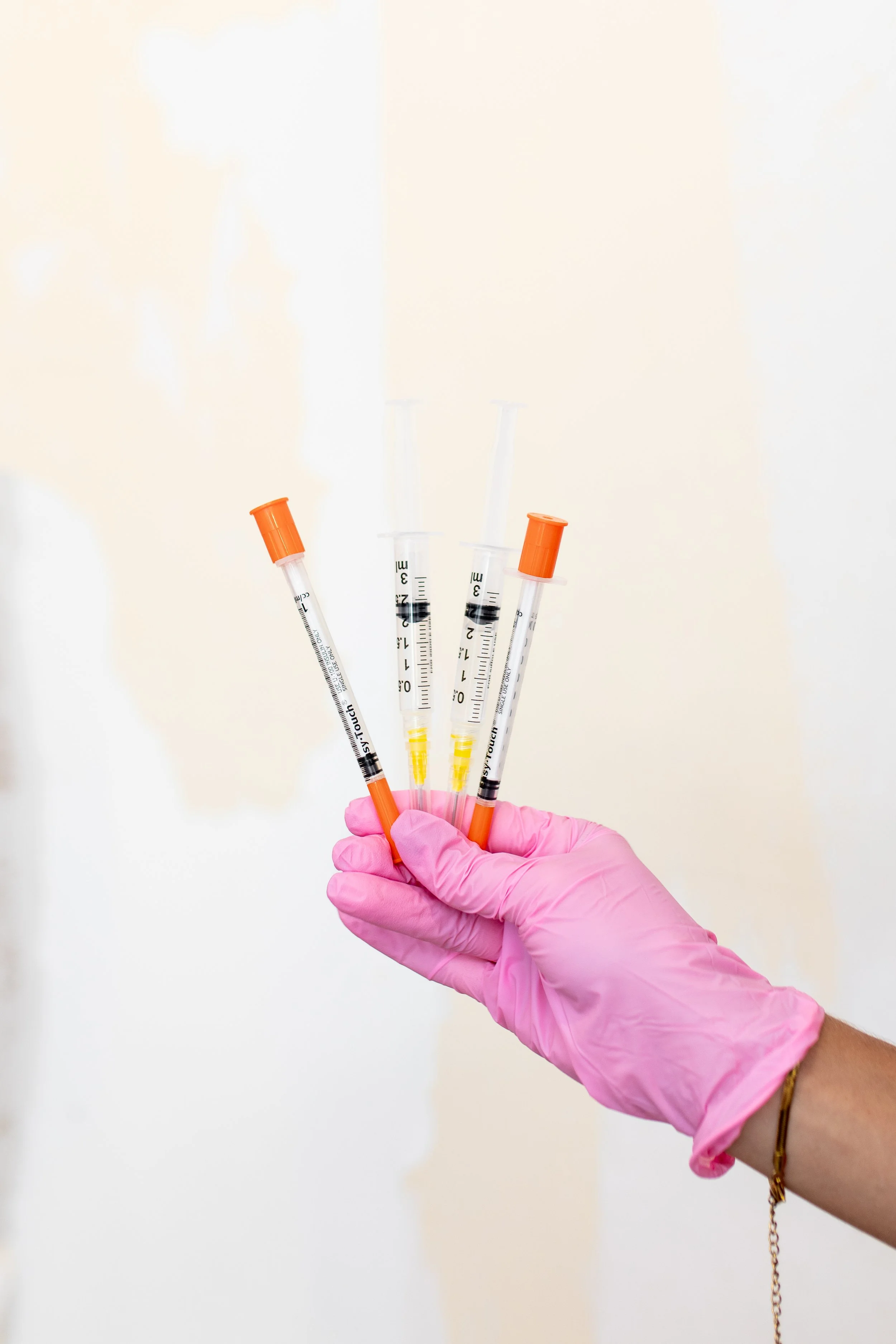 A person's hand wearing a pink glove holding four safety syringes with orange caps and black plungers against a plain background.