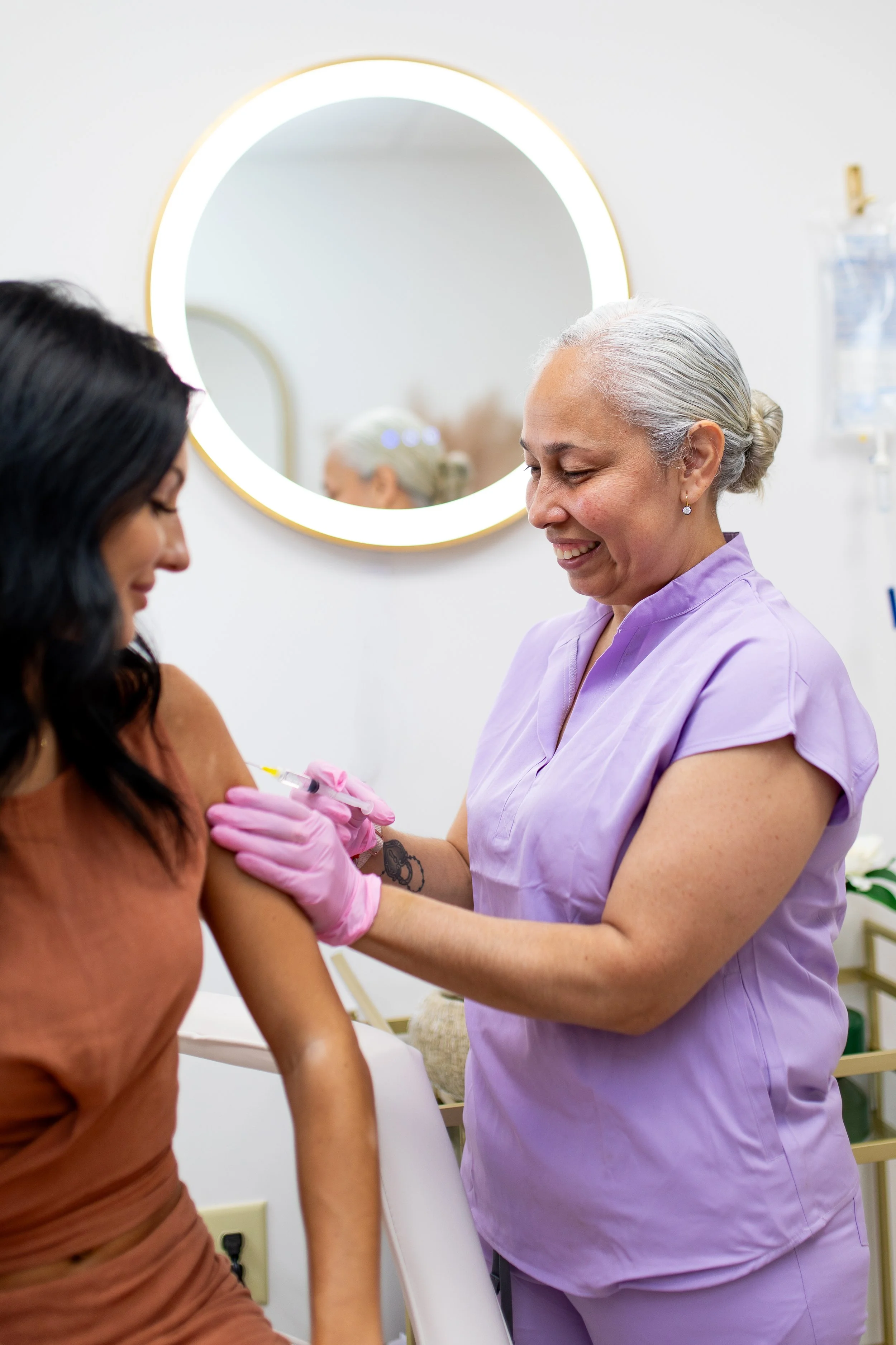 A healthcare worker in purple scrubs administering a vaccination to a woman with dark curly hair, while the woman smiles.