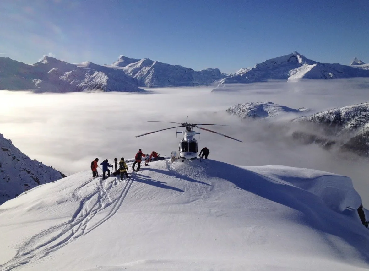 A helicopter on a snow-covered mountain with a group of skiers and a mountain range in the background.