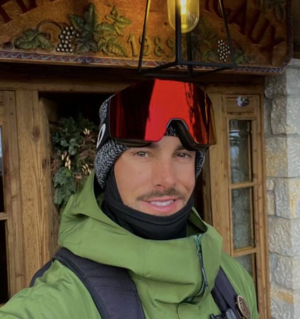 A man in ski gear wearing goggles and a knit hat, smiling for a selfie, with a rustic wooden and stone building in the background in the resort Val Thorens on the 3 Vallées.
