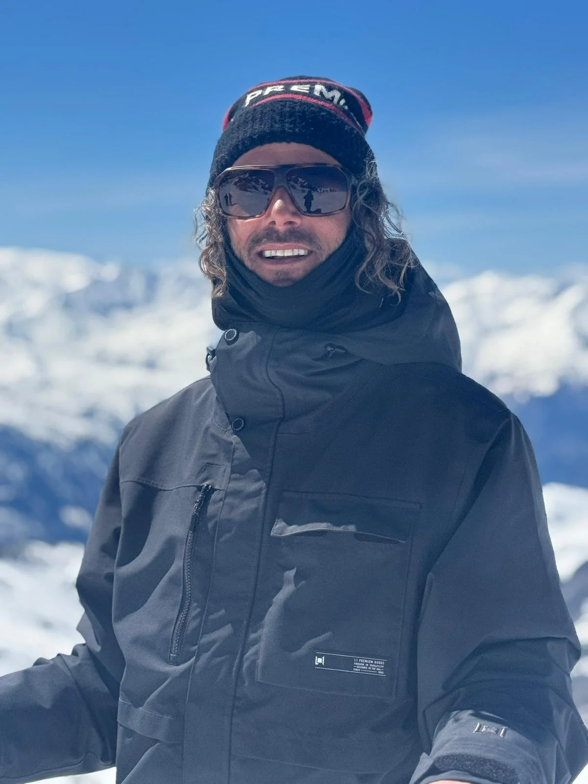 A person wearing a black jacket, a beanie, sunglasses, and a neck gaiter outdoors in a snowy mountain landscape in the resort Val Thorens on the 3 Vallées.