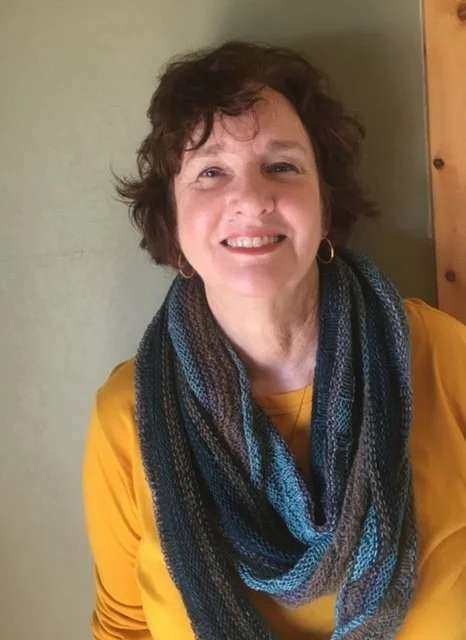 A woman with short curly brown hair, wearing a mustard yellow top and a multicolored blue and gray scarf, smiling indoors against a light green wall and wooden door
