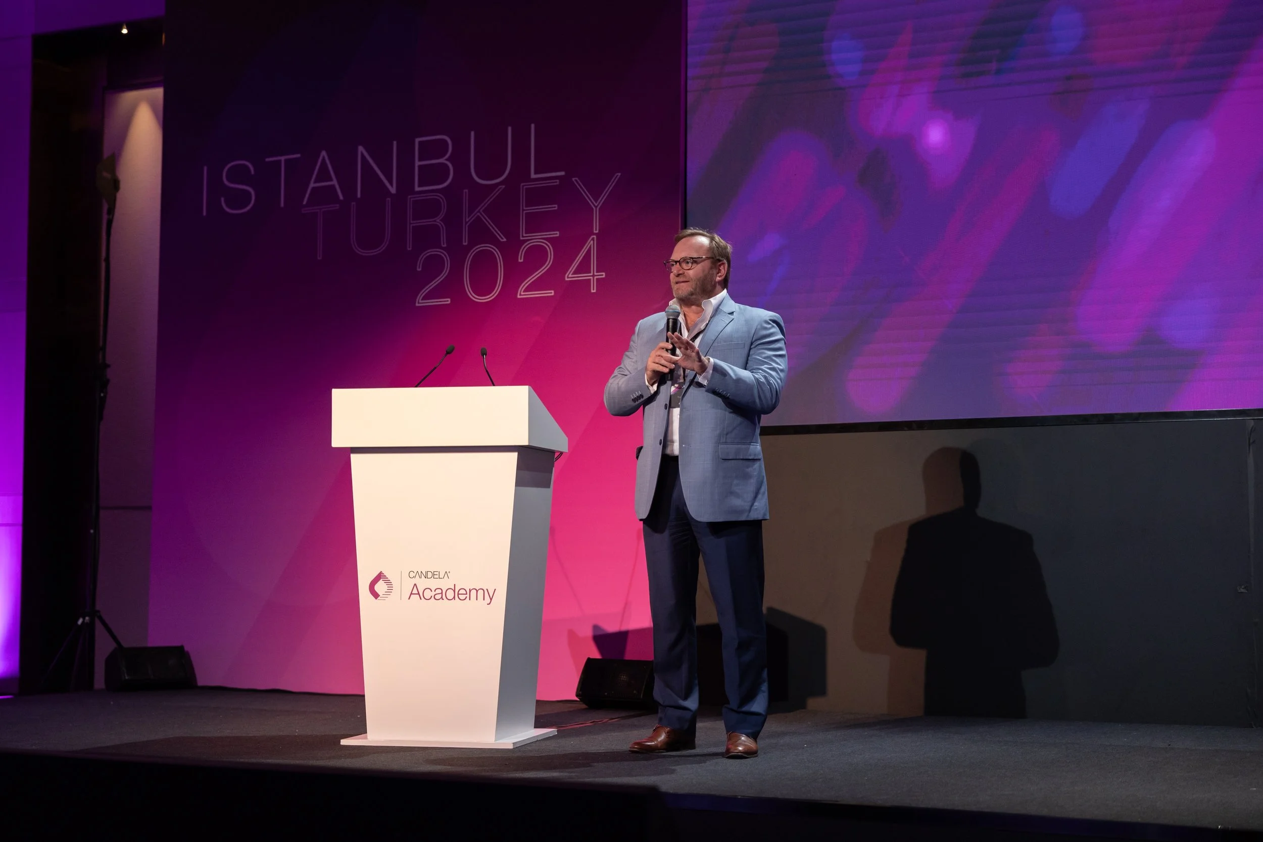 A man in a light blue suit and glasses standing on a stage holding a microphone, with a large screen behind him displaying pink and purple graphics. The stage has a white podium with 'CANDELA Academy' written on it and the background features text re