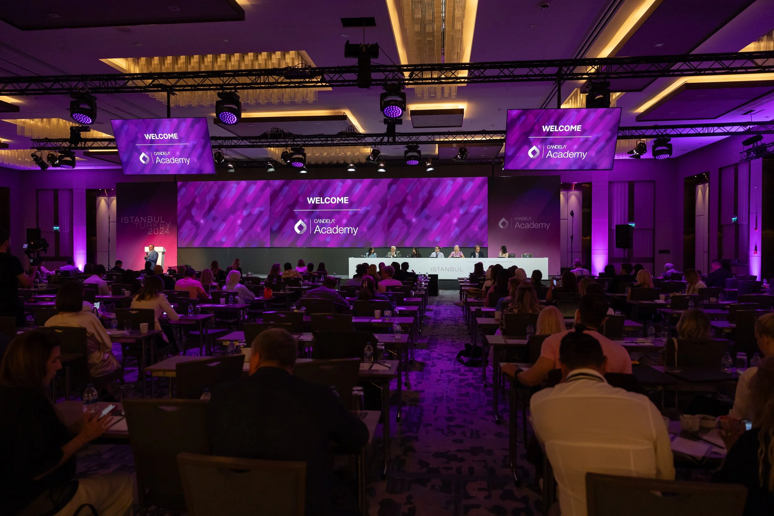 A conference room filled with people seated at tables, facing a stage with a large screen displaying 'Welcome' and 'Candela Academy.' The room has purple lighting, and several smaller screens hang from the ceiling.
