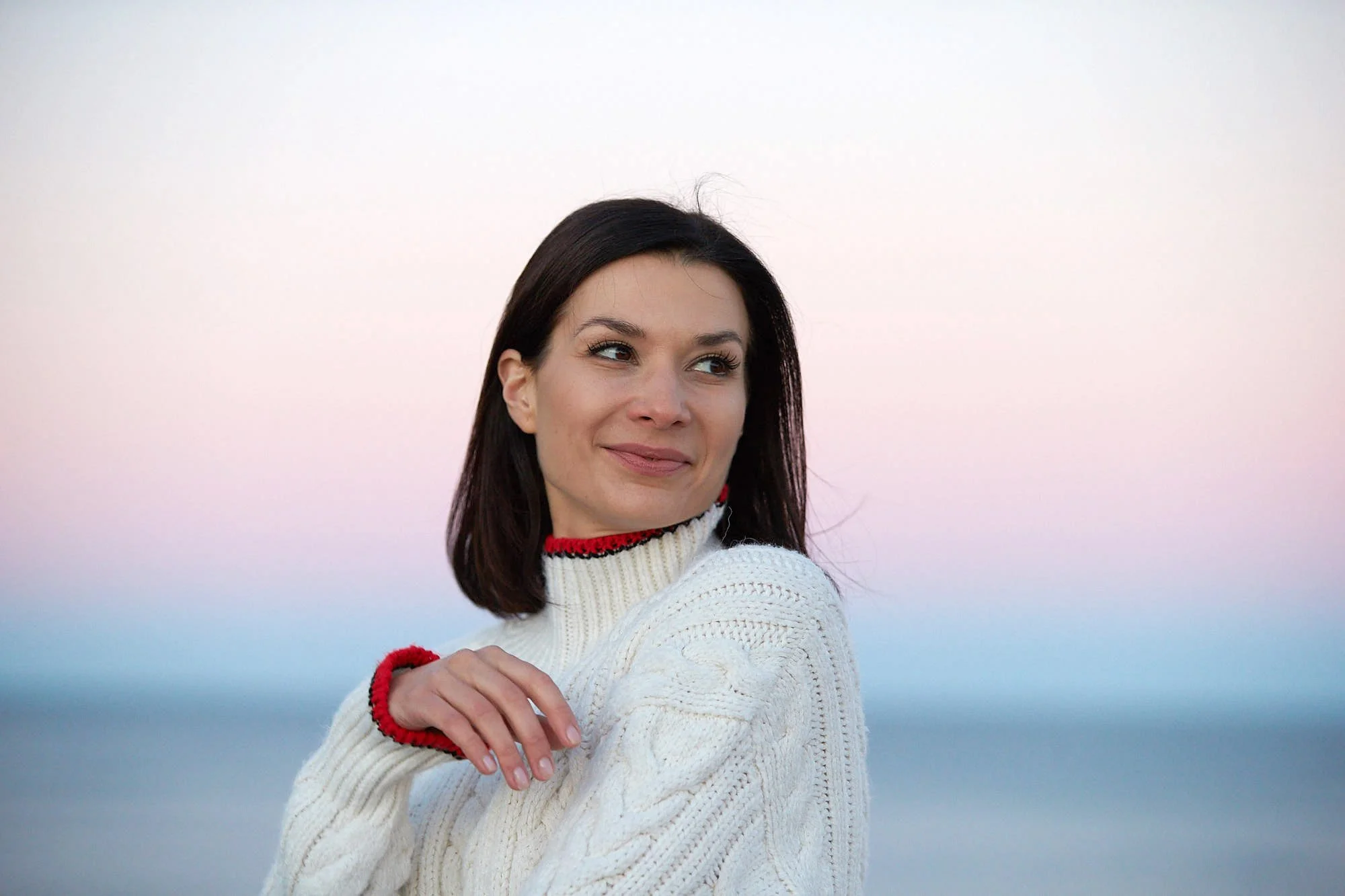 Cynosure brand photography of a woman with dark hair wearing a white knit sweater with red and black accents, smiling and looking to her left, against a pastel-colored sky background.