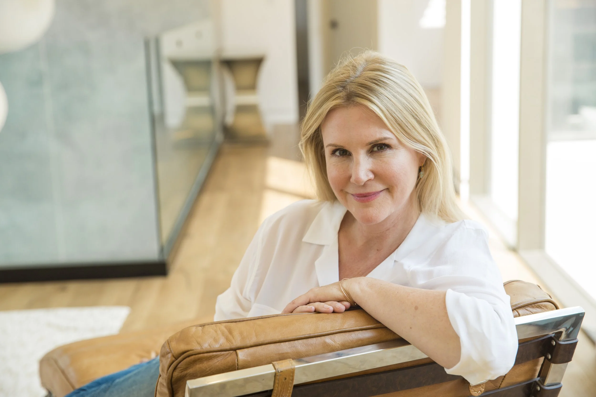 MonaLisa Touch branding photography of a  woman with blonde hair sitting at a wooden table, looking at the camera and smiling in a bright room with large windows.