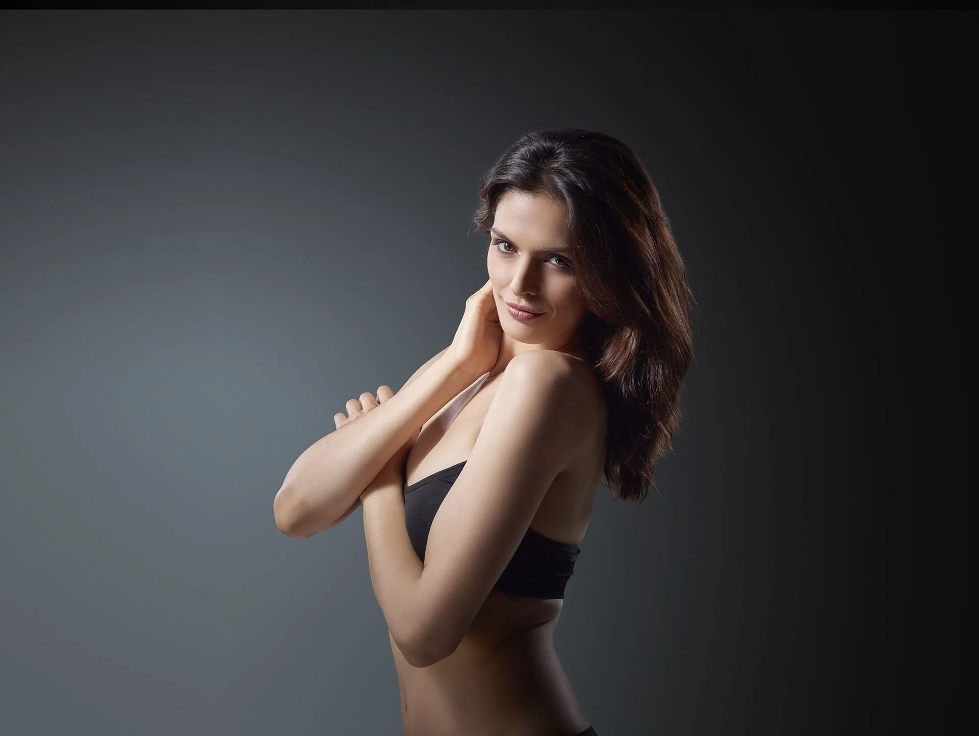 Cynosure brand photography of a woman with brown hair poses with her arms crossed, wearing a black bra, against a dark gradient background.