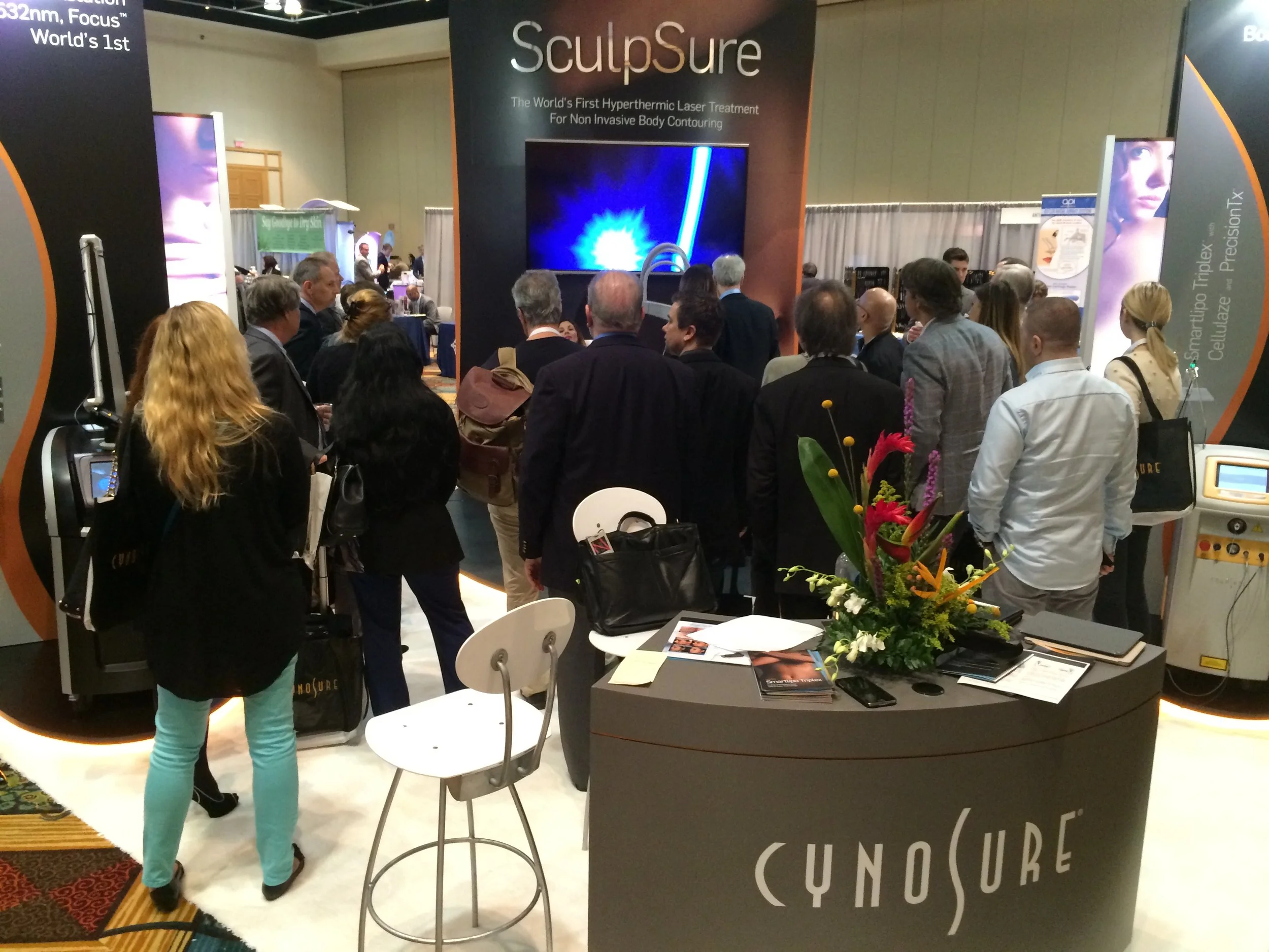 A group of people at a trade show booth for SculpSure, a hyperthermal laser treatment for non-invasive body contouring, with a large screen, informational posters, and a table with magazines, brochures, and a flower arrangement.