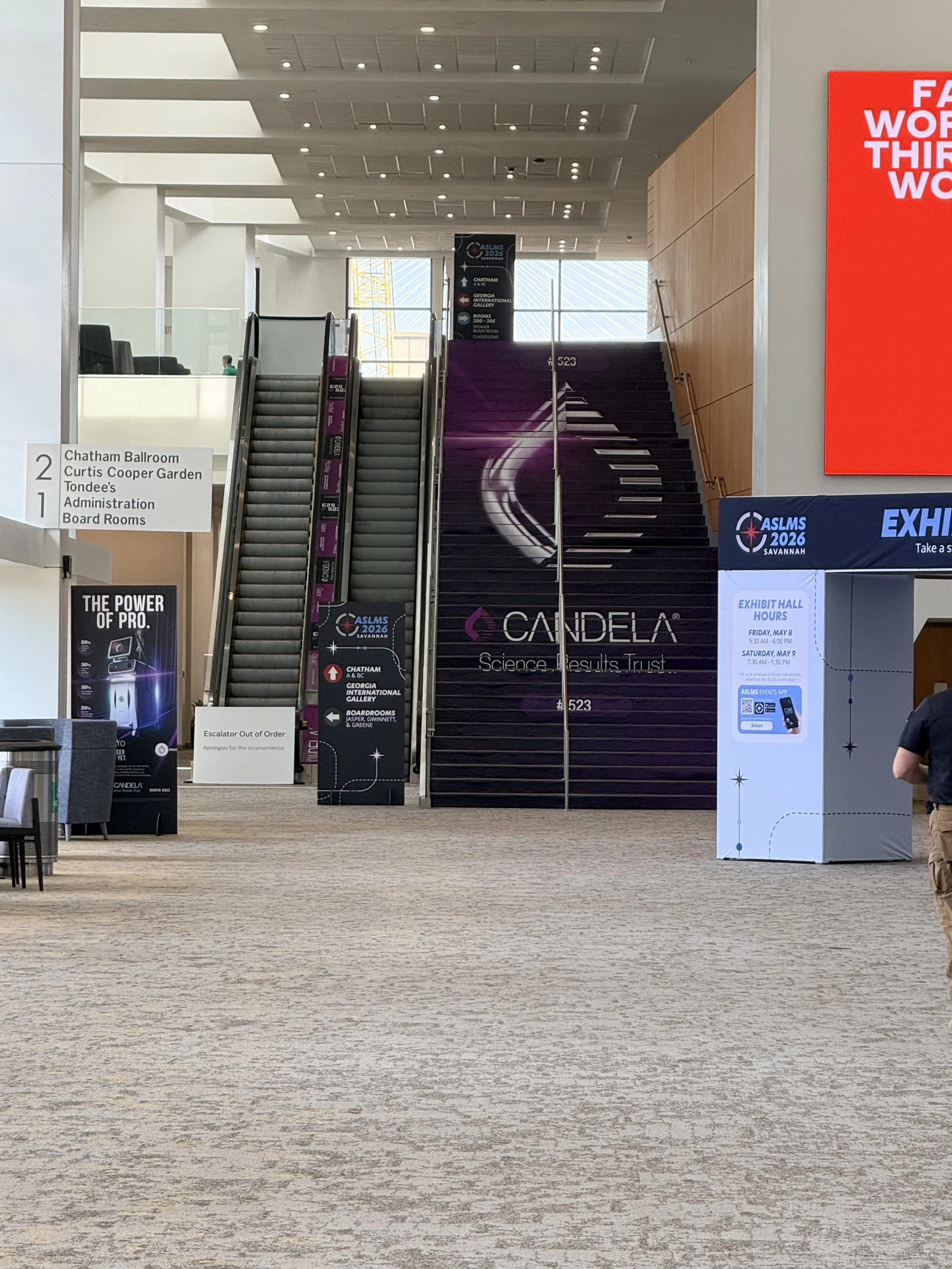 Interior view of a convention center with escalators, signage for ASLMS 2026, and banners for events. The floor is carpeted in beige, and there are a few chairs with black seats and silver frames. Large sky windows let in natural light.