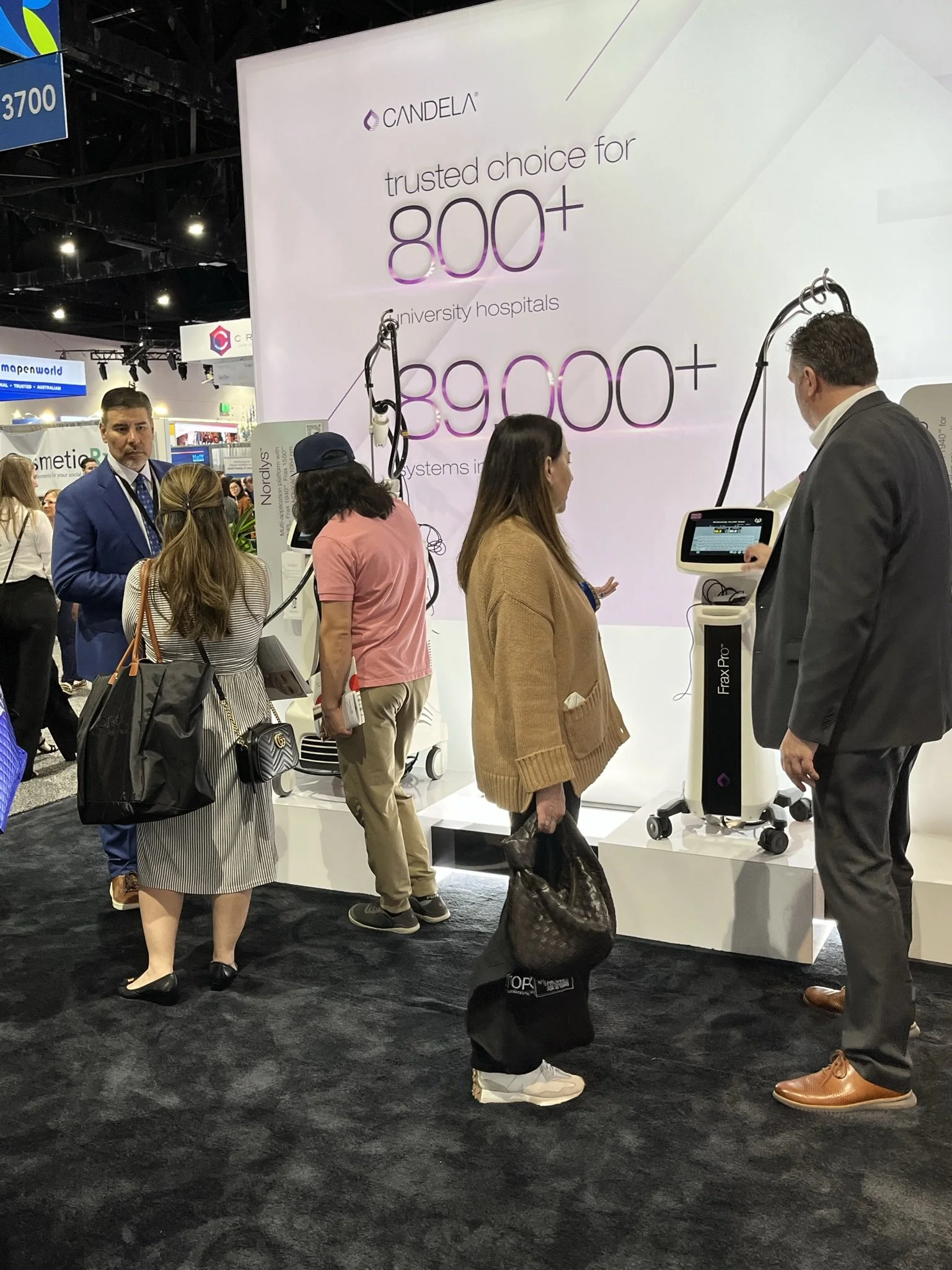 People at a trade show or exhibition, gathered around medical or technological equipment, with a large sign behind them displaying the text 'trusted choice for 800+ university hospitals.'