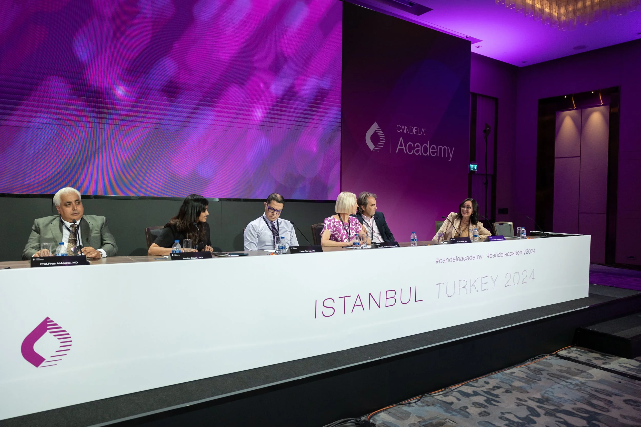 A panel of six people sitting at a long table on a stage during a conference or panel discussion at the Candela Academy event in Istanbul, Turkey, 2024. The background features a large purple and pink digital display and the Candela Academy logo.
