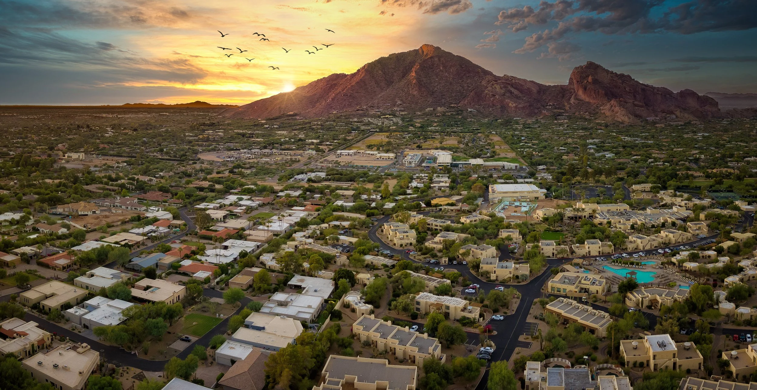 Aerial view of a residential area with houses, roads, and trees, with a mountain in the background during sunset, and birds flying in the sky.