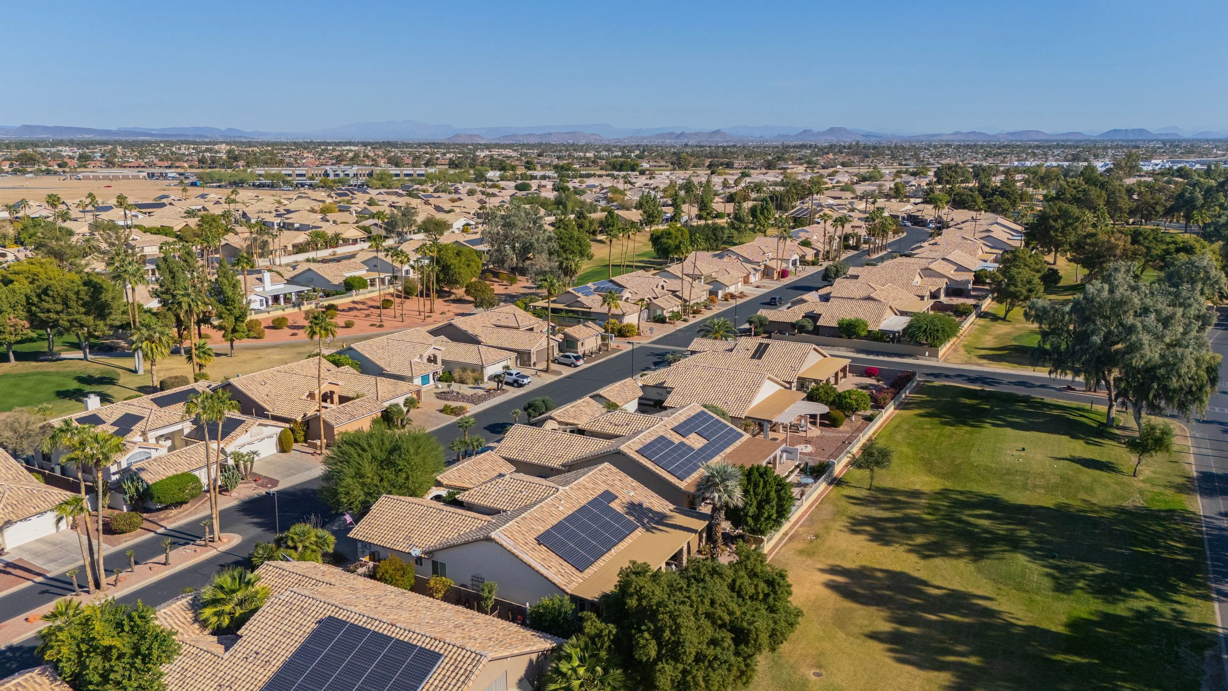 Aerial view of a suburban neighborhood with single-story houses, tiled roofs, and solar panels, surrounded by trees and green lawns.