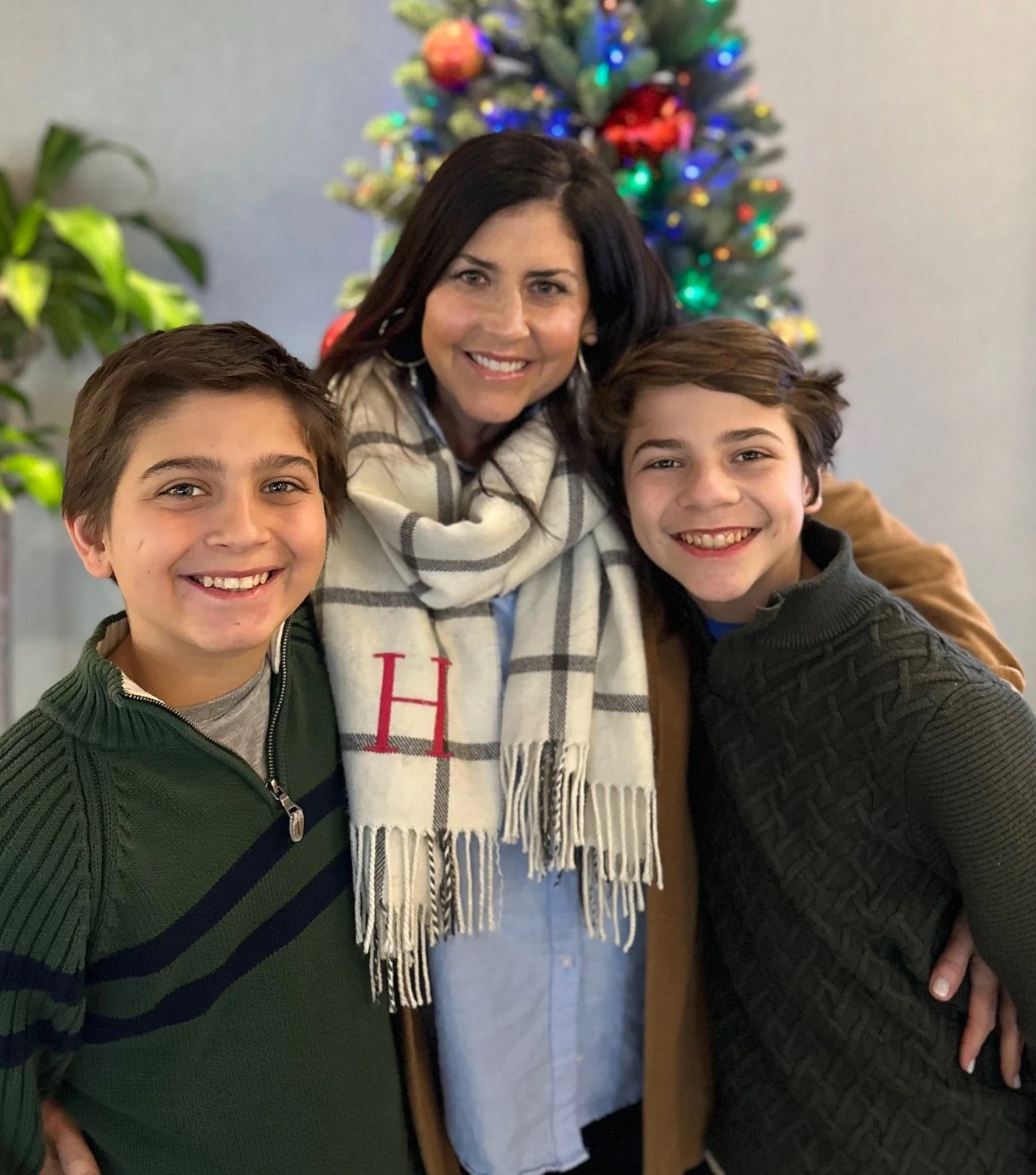 A woman and two boys smiling in front of a decorated Christmas tree, with a plant visible on the side.