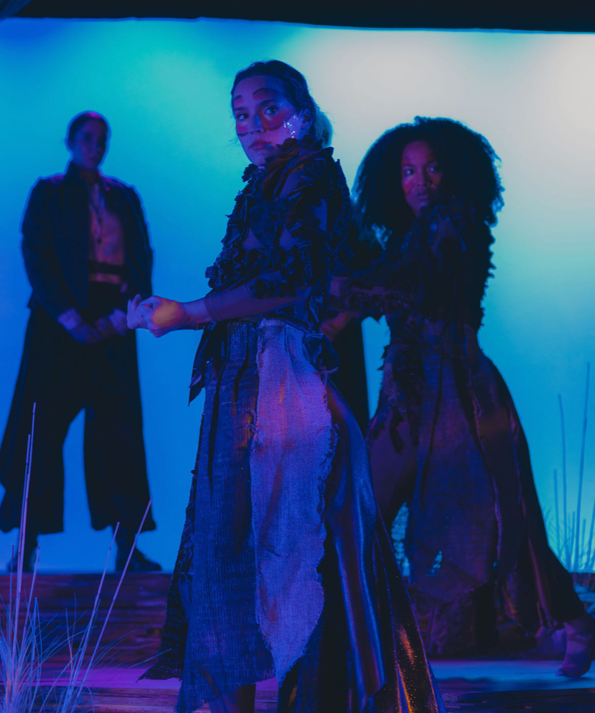 Three women in dramatic, dark clothing on a stage with blue lighting, one woman front and center looking directly at the camera, two in the background.