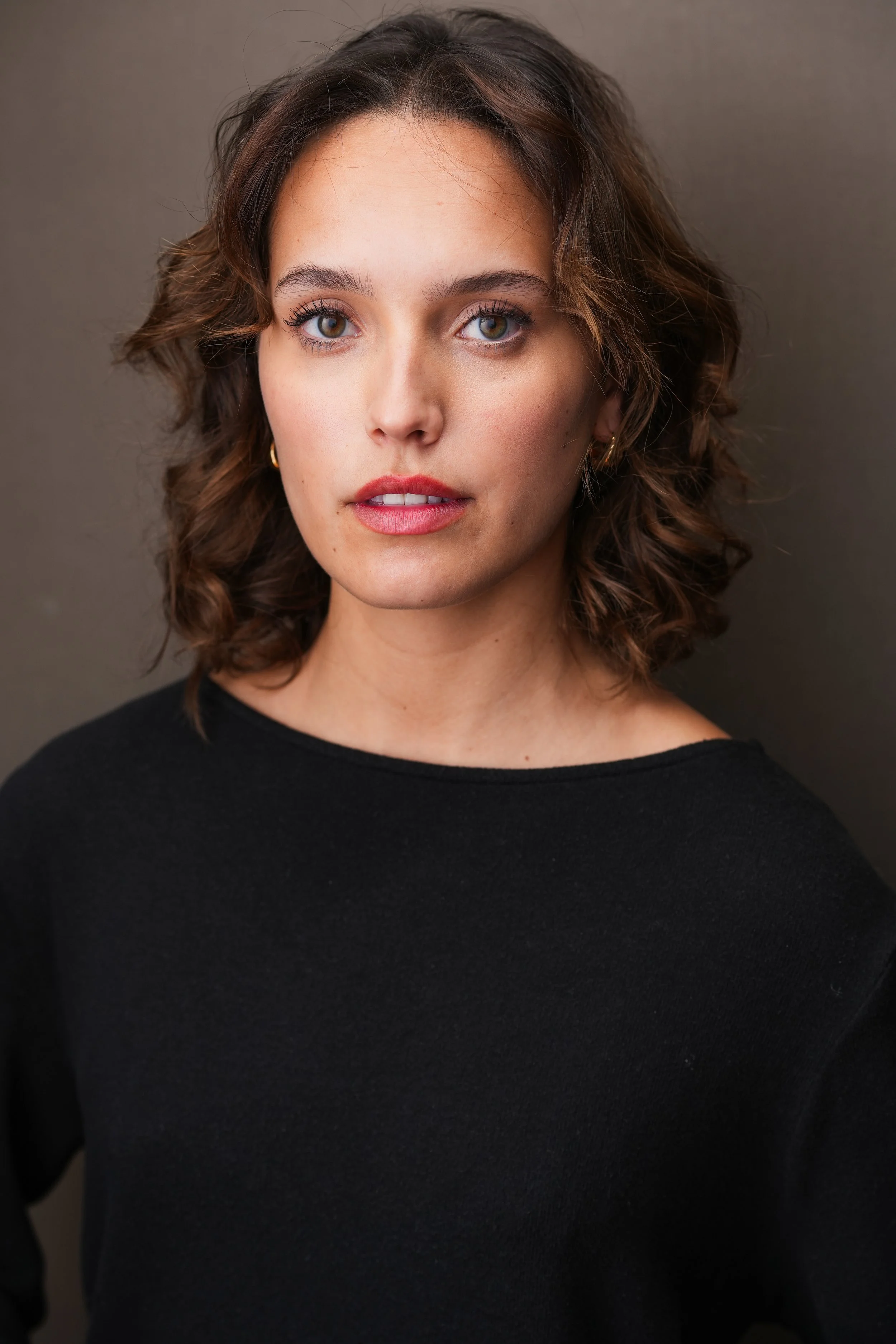 Close-up of a woman with wavy brown hair, blue eyes, and light makeup, wearing a black top, against a neutral background.