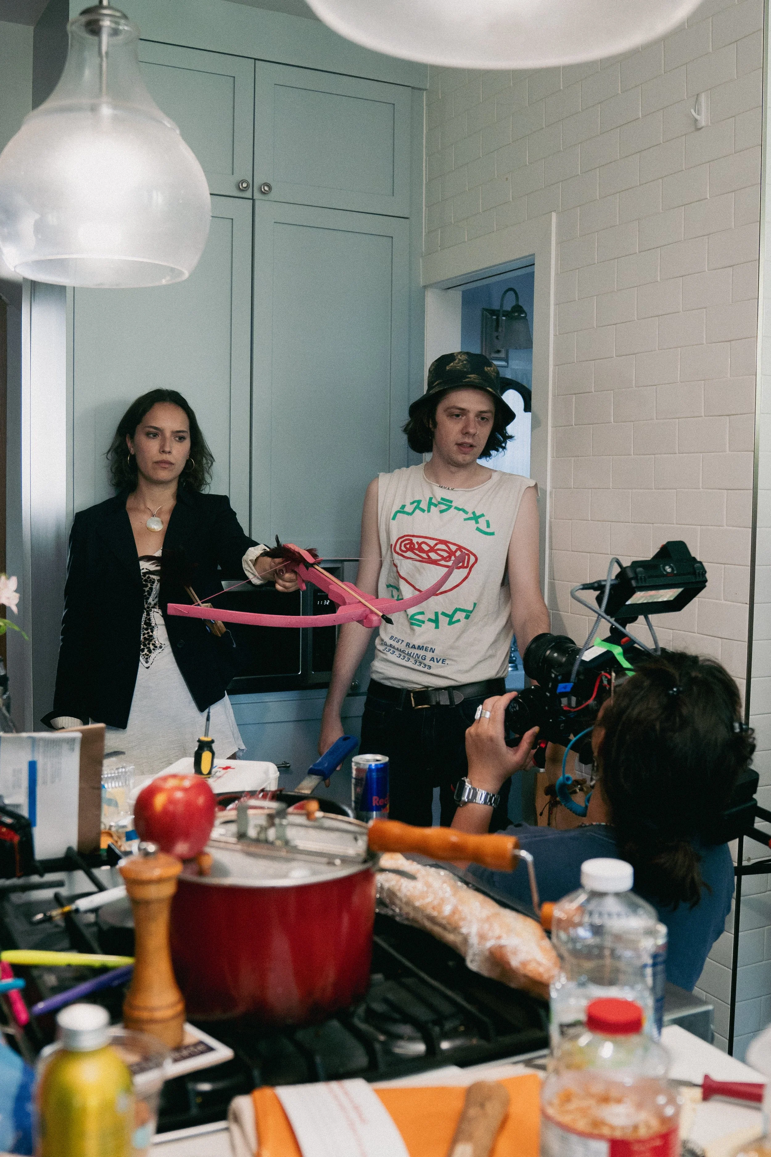 Two women standing in a kitchen behind a counter with various food items and utensils. The woman on the left has dark hair, wears a black jacket and a white dress, and is holding a kitchen utensil. The woman on the right has shoulder-length dark hair