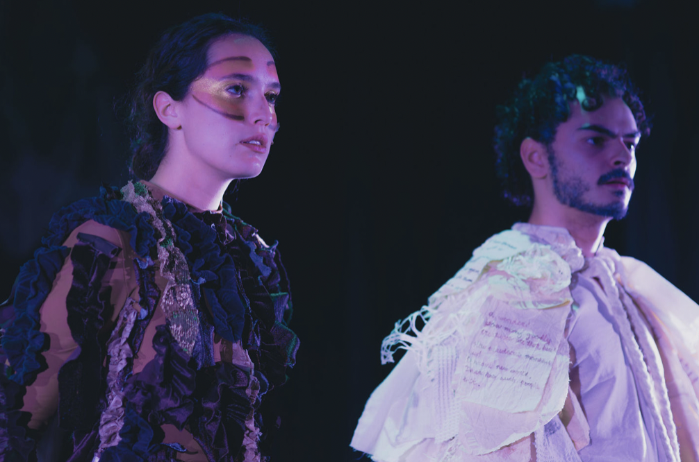 Two models standing side by side in dark setting, dressed in elaborate, textured clothing with dramatic lighting.