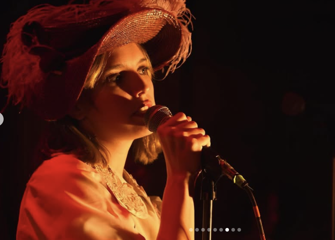 A woman wearing a large, wide-brimmed straw hat with fringed edges, holding a microphone and singing or speaking on stage with warm red lighting.