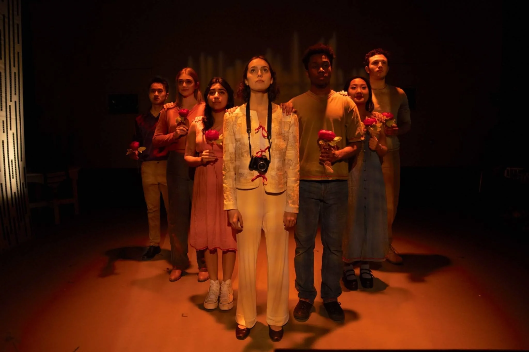 Six young adults standing in a dark room, each holding a pink rose, with the central woman also holding a camera around her neck, positioned under a dim spotlight.
