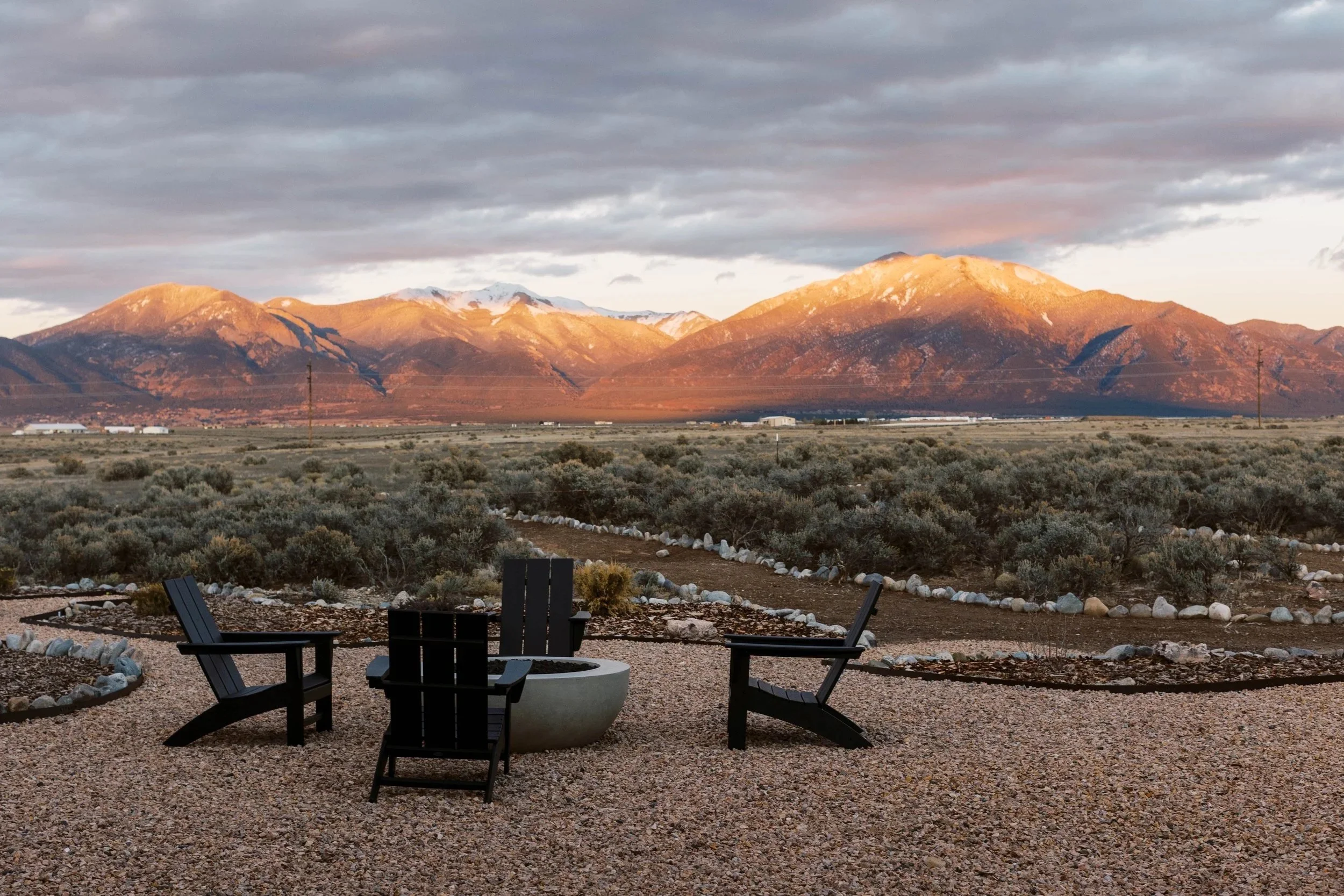Outdoor seating area with black chairs and a fire pit, overlooking desert landscape with mountains in the background and cloudy sky.