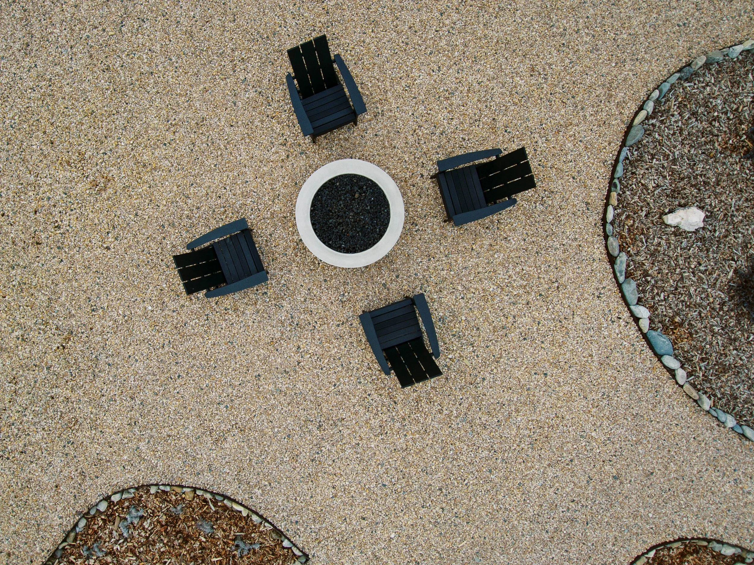 Overhead view of a sandy outdoor area with four black Adirondack chairs arranged around a white fire pit filled with black gravel, bordered by a curved border of rocks and mulch.
