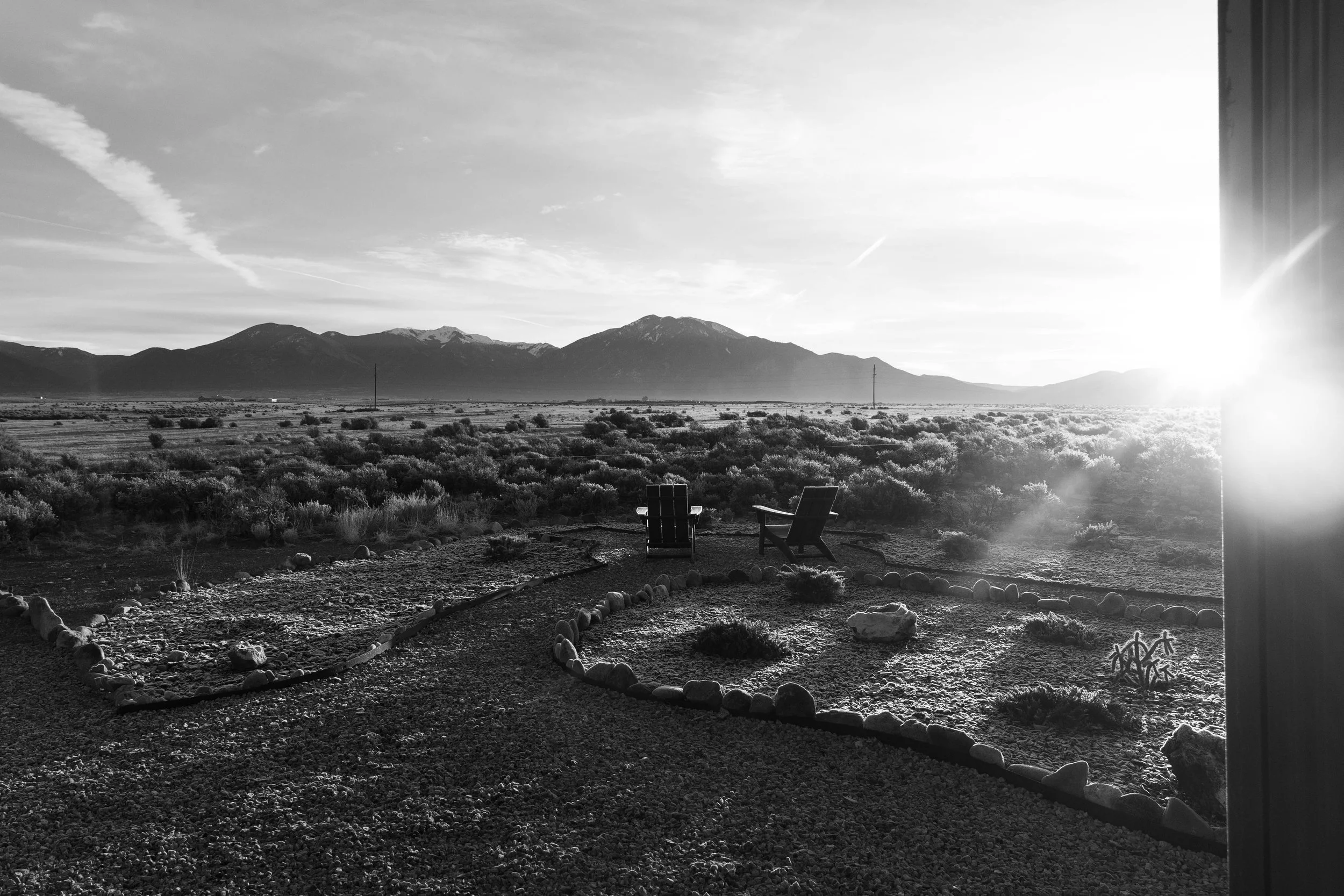 Black and white photo of a desert landscape with mountains in the background, a field of shrubbery, and two chairs near a round garden bed, with a partially visible window on the right side.