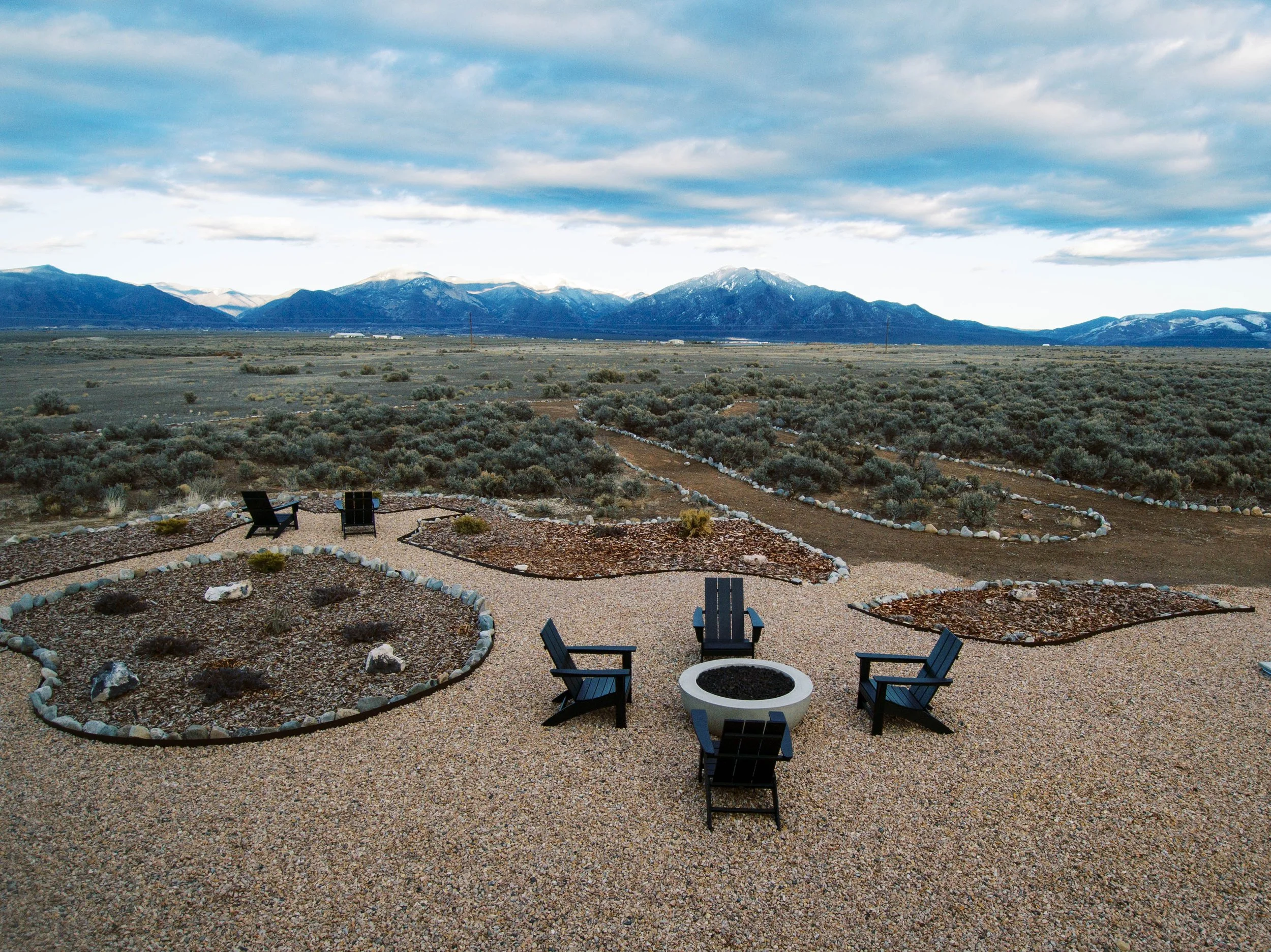 Outdoor seating area with black chairs around a fire pit, gravel ground, desert plants, and mountain landscape in the background under a partly cloudy sky.