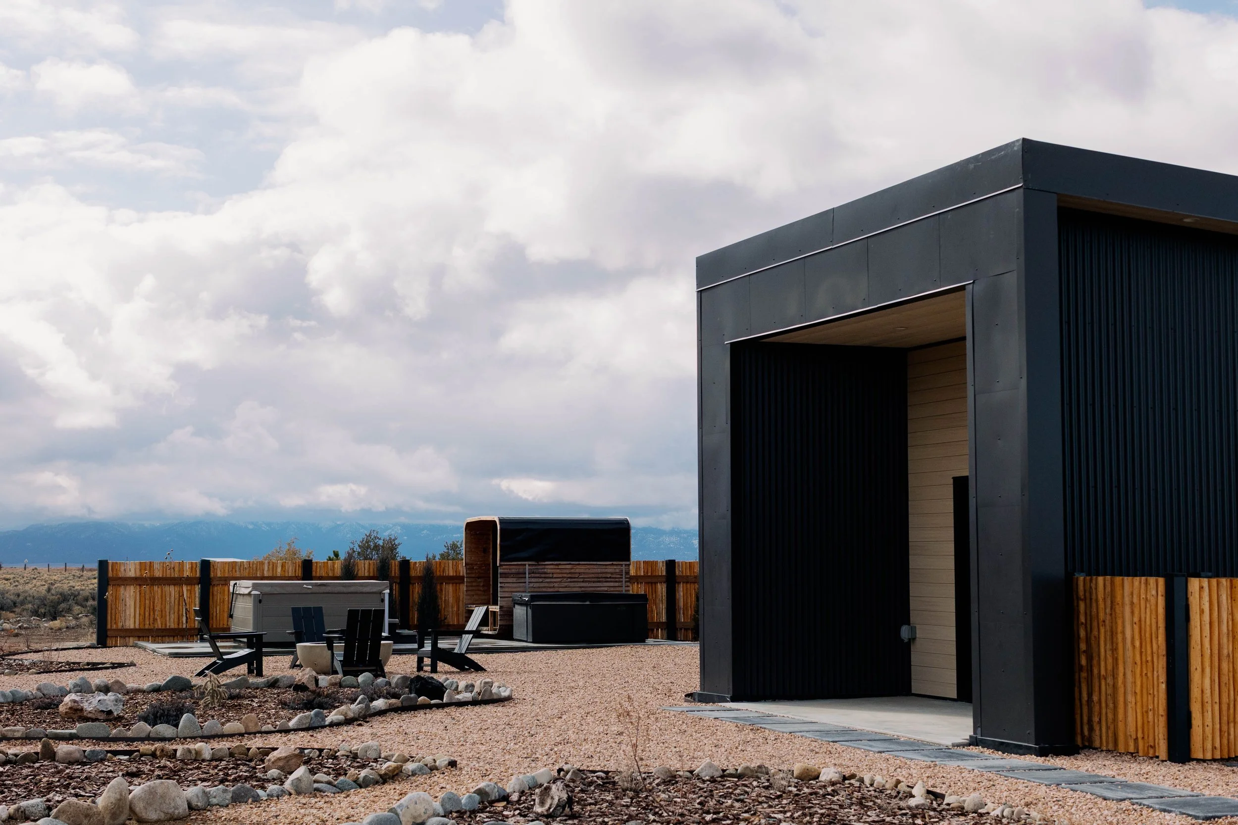 Part of a modern house exterior with a dark gray wall and wooden accents, outdoor seating area with chairs and tables, a wooden fence, and mountains in the background under a partly cloudy sky.