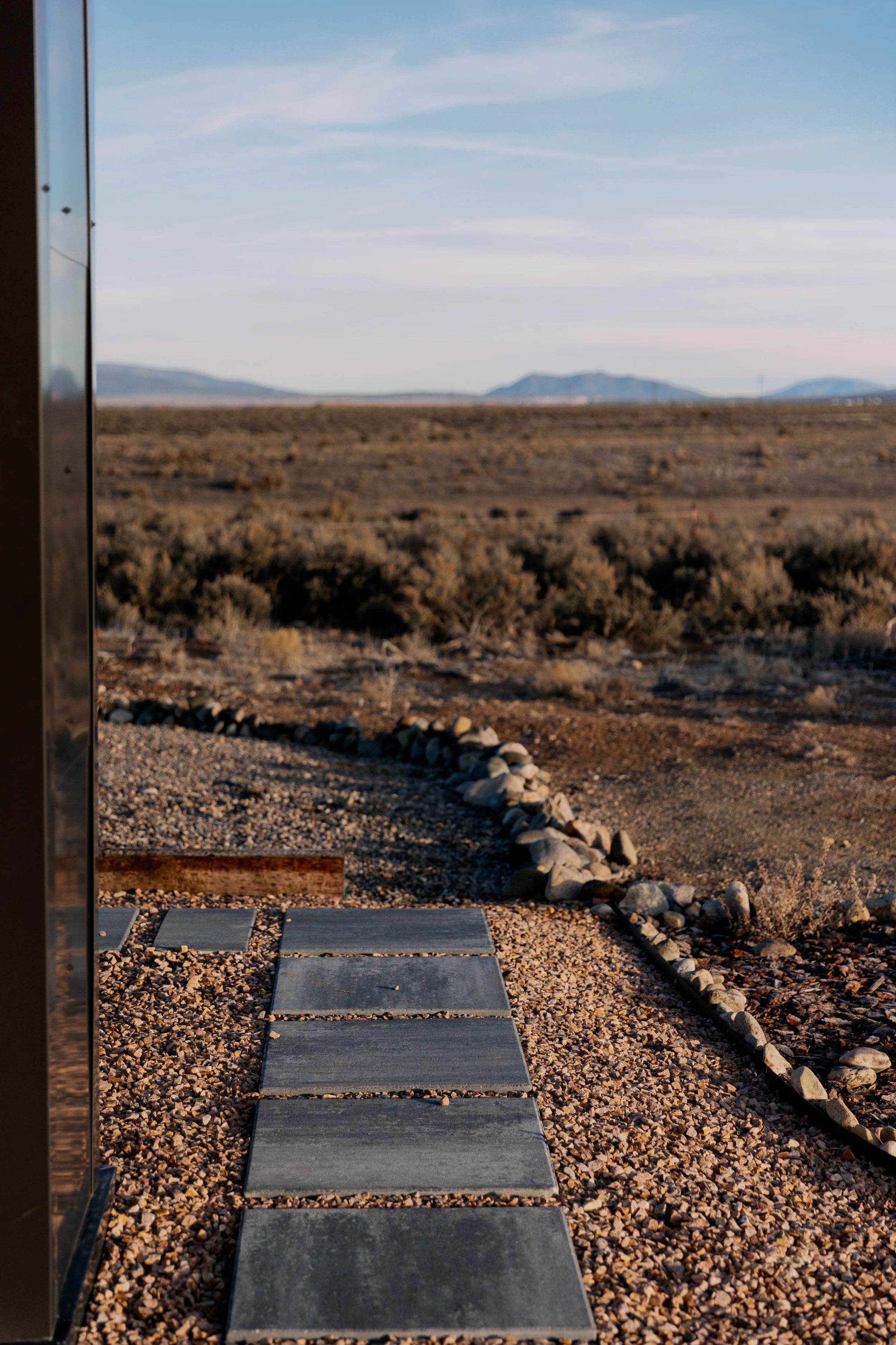 Desert landscape with mountain range in the distance, blue sky with scattered clouds, and a pathway made of rectangular stone slabs on gravel. Rocks border the pathway, and a metal structure is visible on the left side of the image.