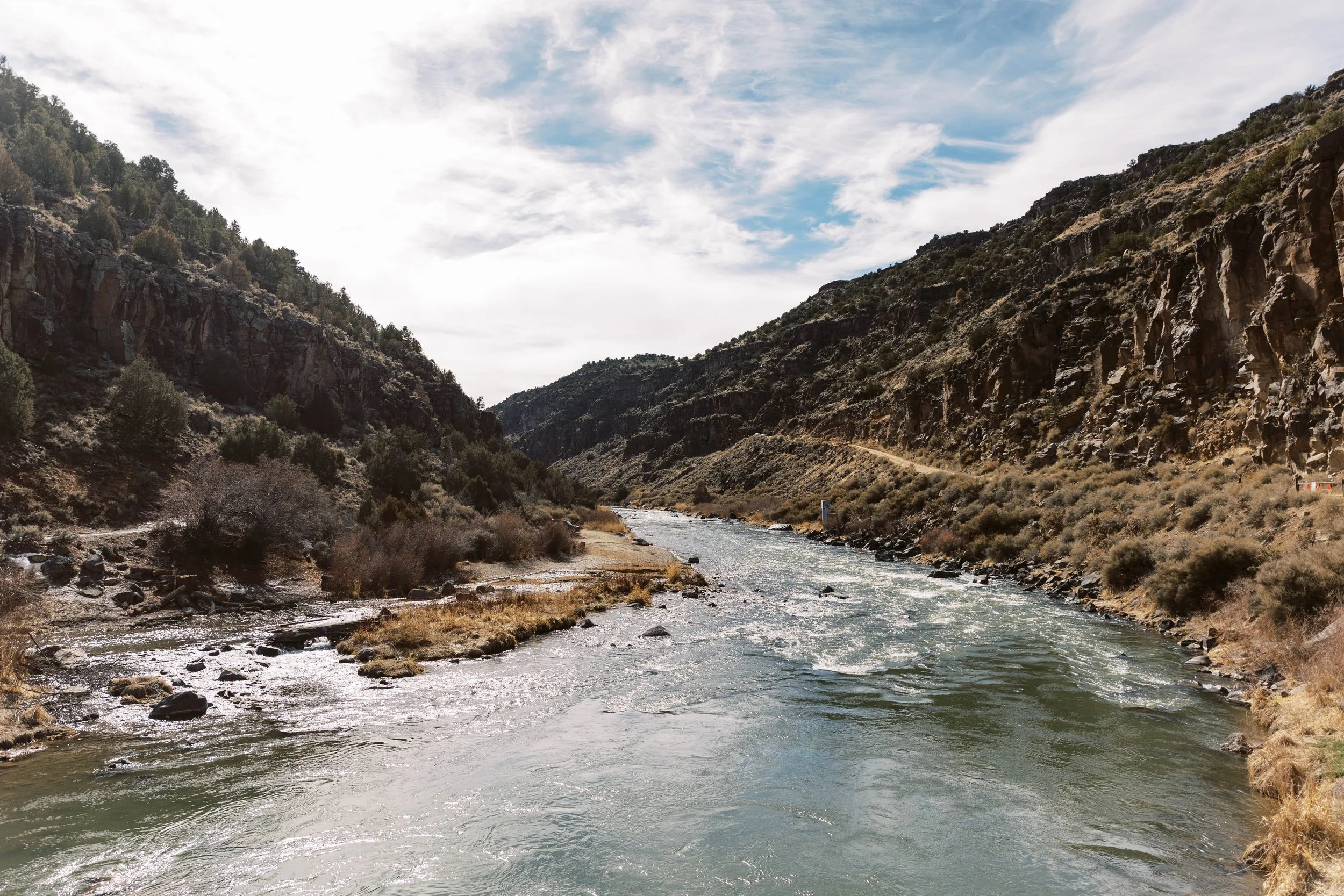 A river flowing through a narrow canyon with rocky cliffs and sparse vegetation under a partly cloudy sky.